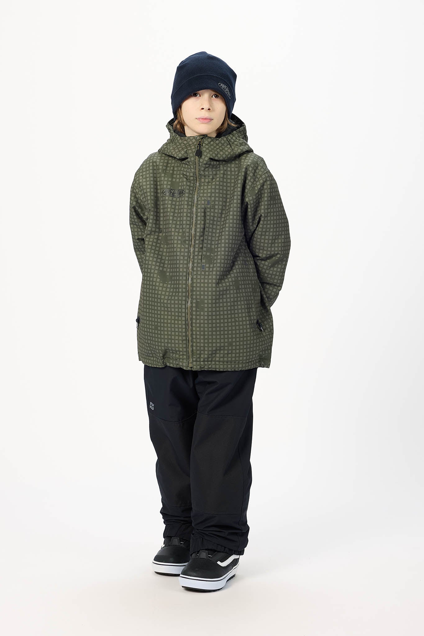 A child model wearing an olive green and black checkered hooded jacket, black pants, a black beanie, and black sneakers, standing against a plain white studio background.