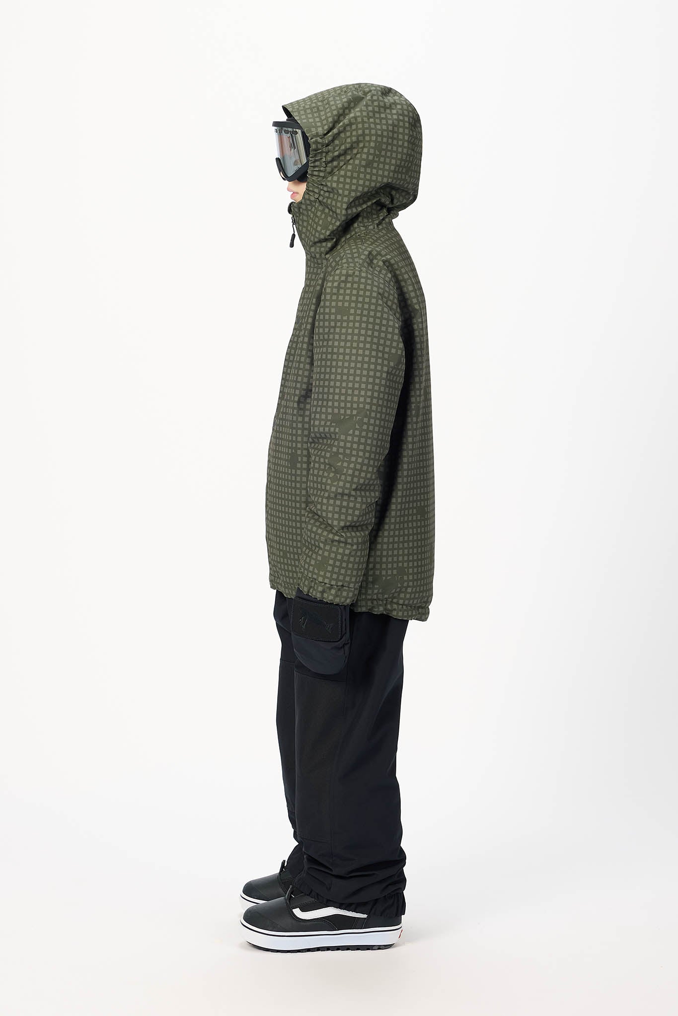 A person shown in profile wearing an olive green hooded jacket with a subtle grid pattern, black pants, and black-and-white sneakers against a plain white studio background.