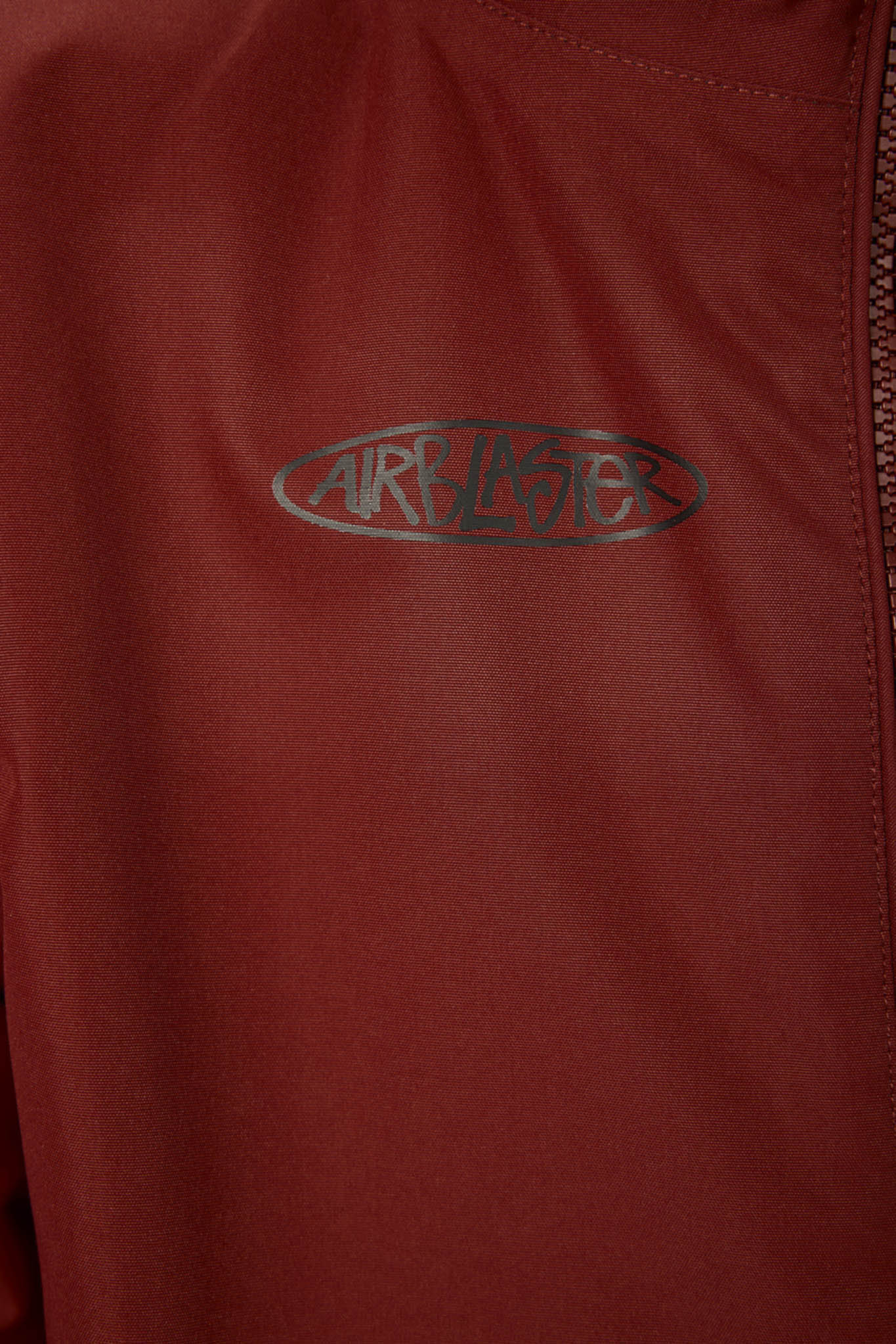 Close-up of a dark red or burgundy jacket with a dark gray metallic Airblaster logo on the chest and a zipper running vertically on the right side.