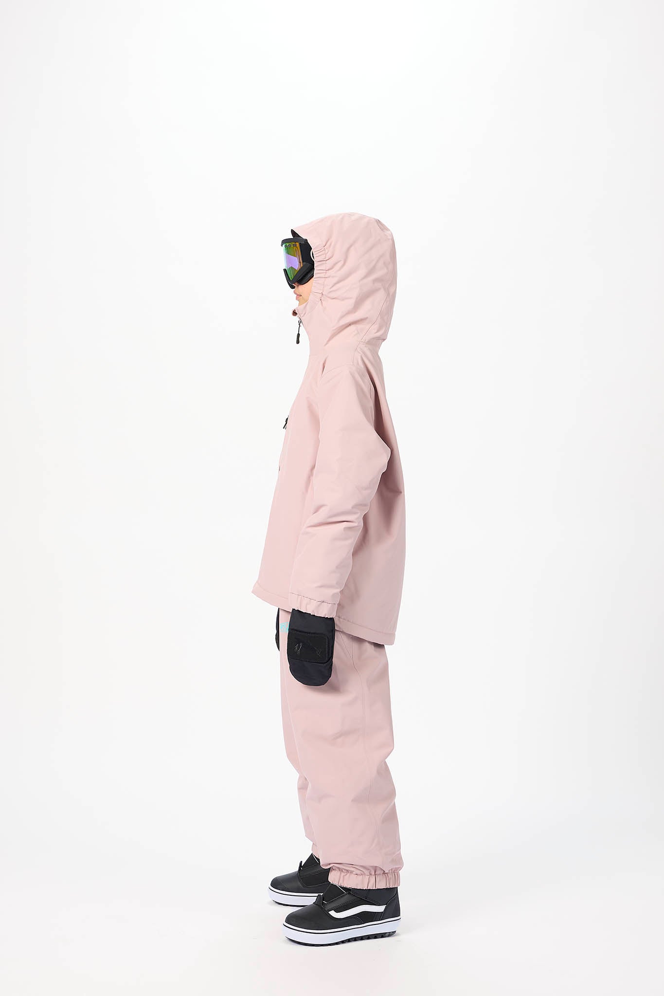 A child standing in profile wearing a light pink hooded snow jacket and matching pants, with black gloves and black sneakers, against a plain white studio background.