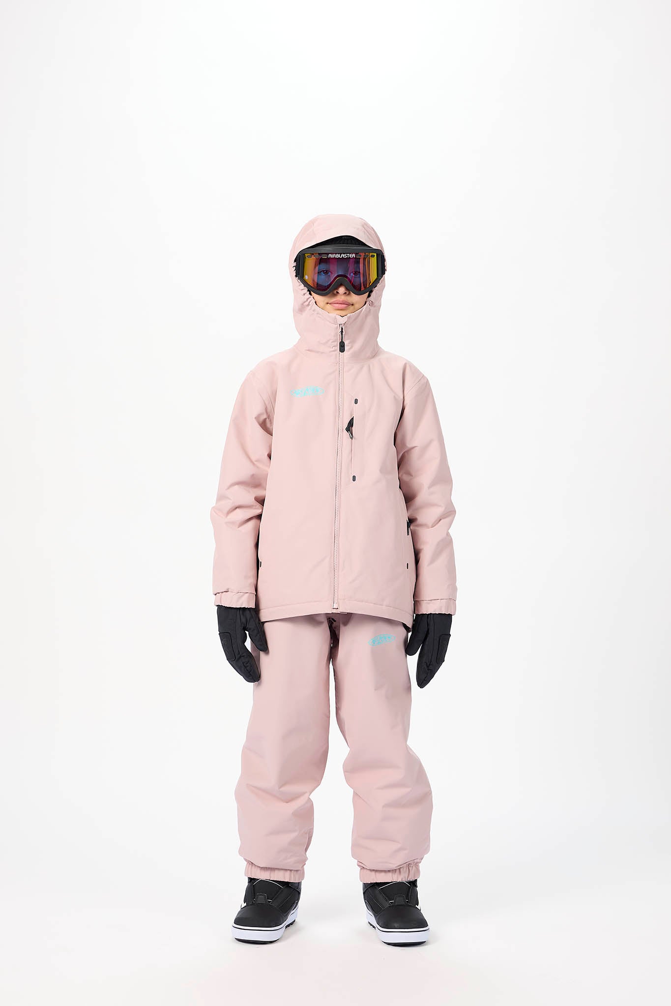 A child standing against a white studio background wearing a light pink snowboard jacket and matching pants, with black gloves and black-and-white snow boots. The child is also wearing ski goggles.