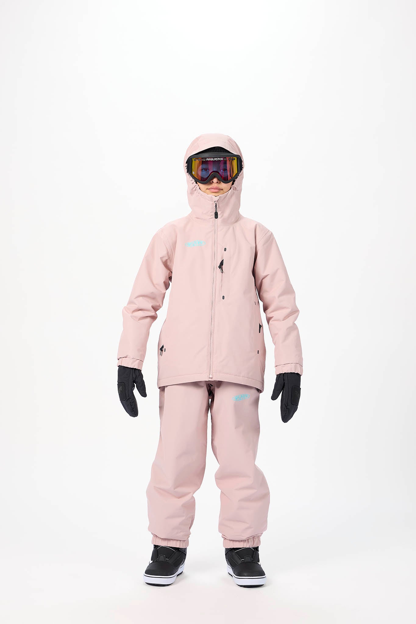 A child wearing a light pink ski jacket and matching pants, with black gloves and ski boots, standing against a plain white studio background.
