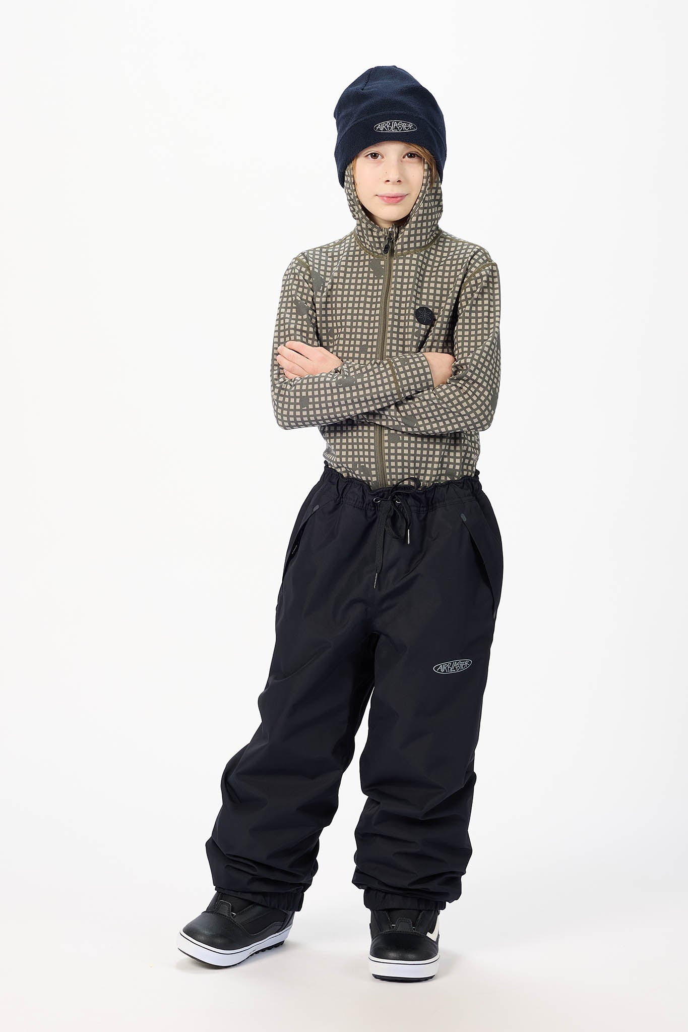 A child model wearing a patterned zip-up jacket, black snow pants, a dark navy beanie, and black sneakers, standing against a plain white studio background.