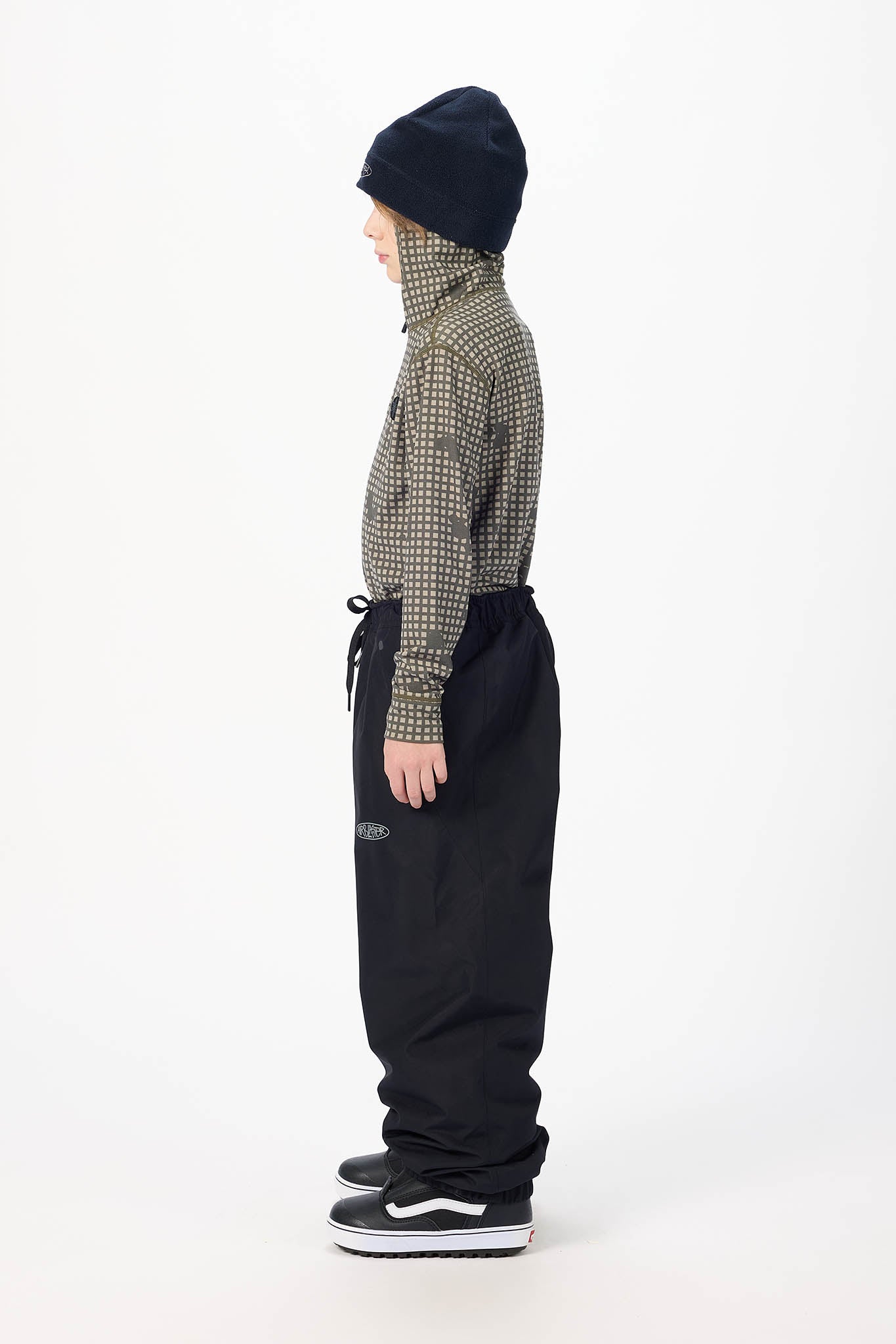 A child model shown in profile wearing a dark navy knit beanie, a green and beige patterned long-sleeve top, black drawstring sweatpants, and black sneakers with white soles, photographed against a clean white studio background.