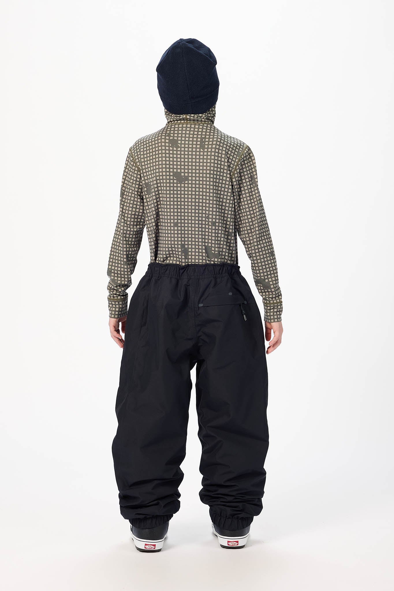 A model shown from the back wearing a patterned long-sleeve shirt, black snow pants, a black beanie, and black sneakers against a plain white studio background.