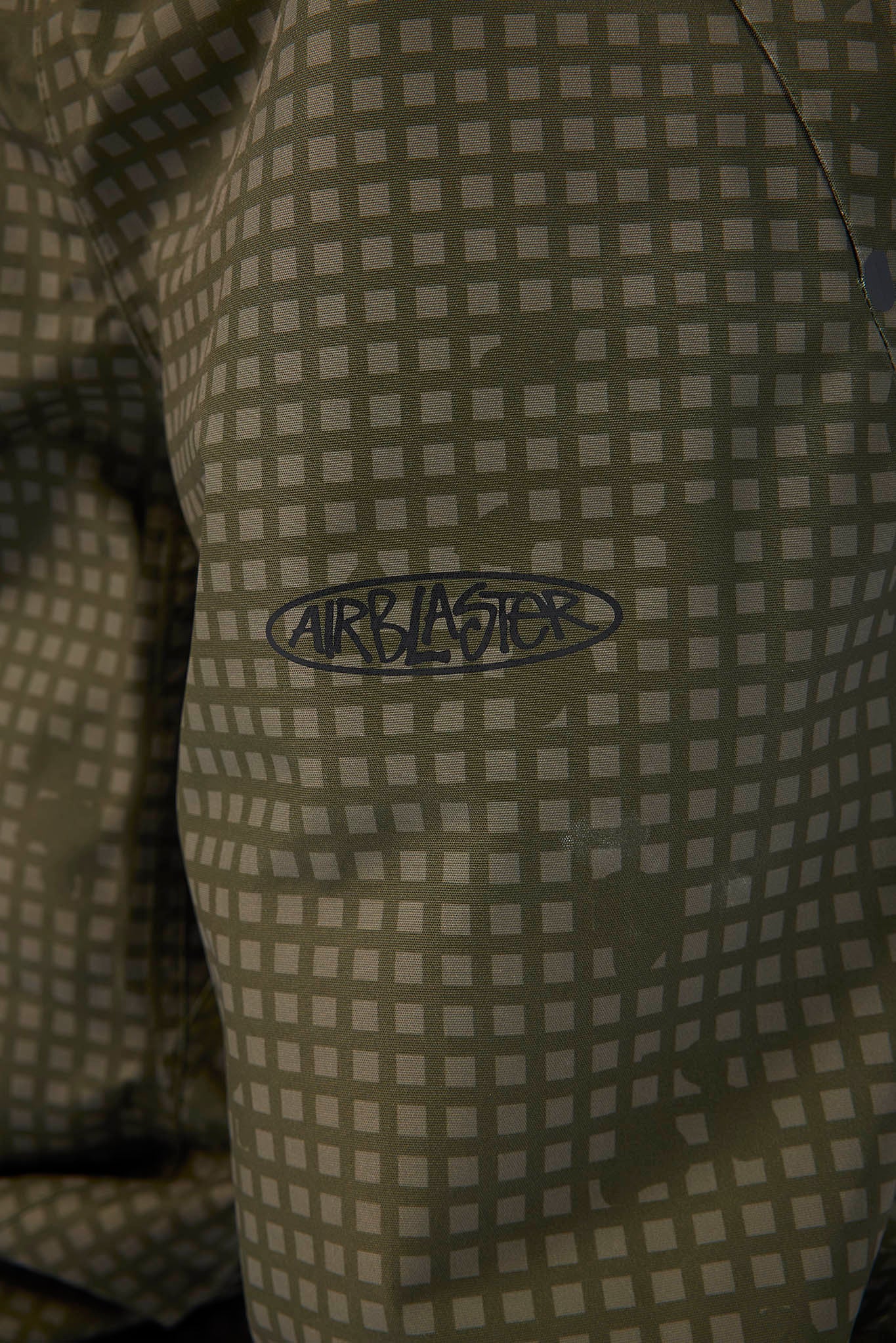 Close-up of a patterned garment featuring a gray and olive green checkered design with a dark gray Airblaster logo embroidered on the chest.