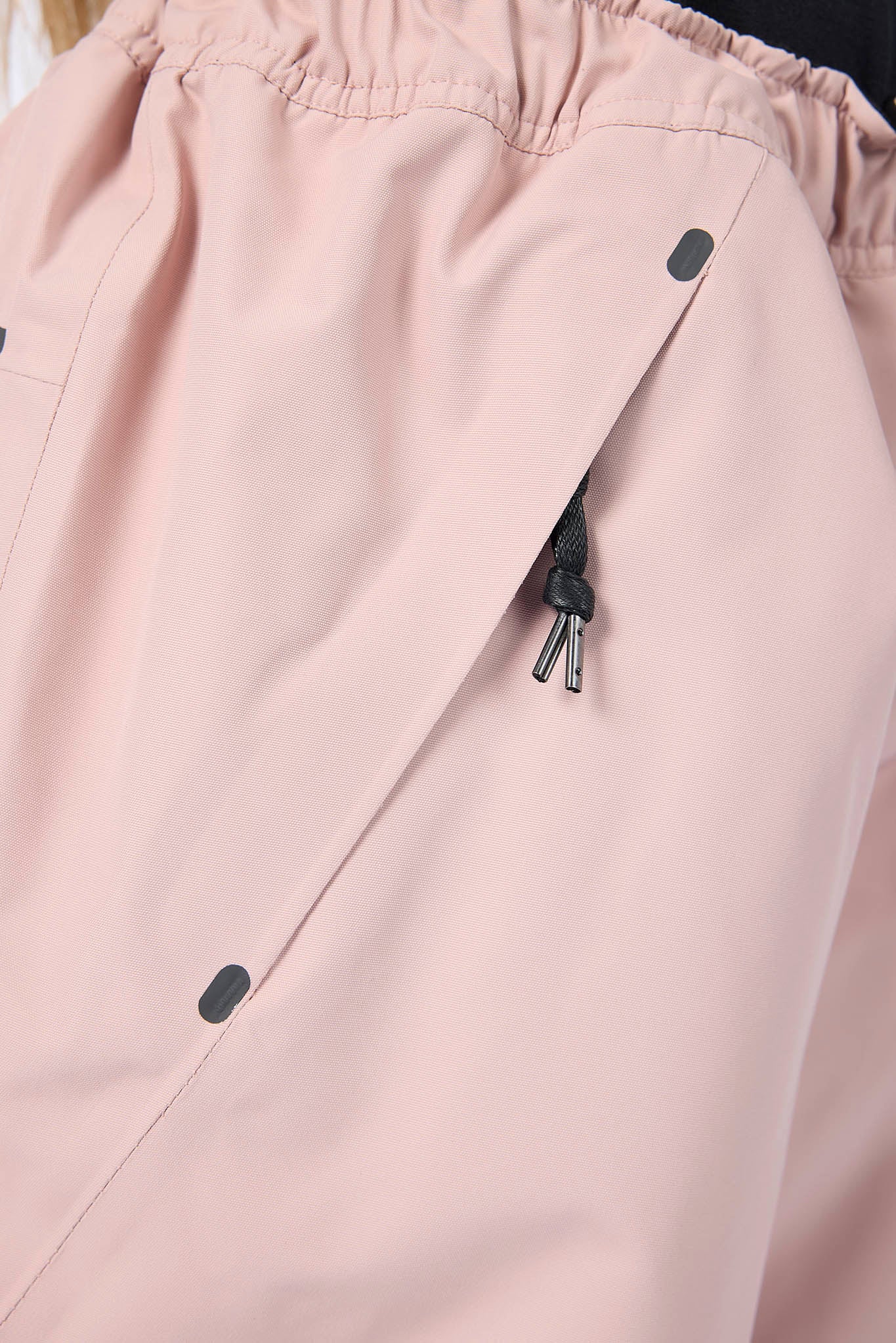 Close-up of a light blush pink garment, likely pants or a jacket, showing an elastic waistband, a diagonal front seam, dark snap buttons, and a black zipper pull.