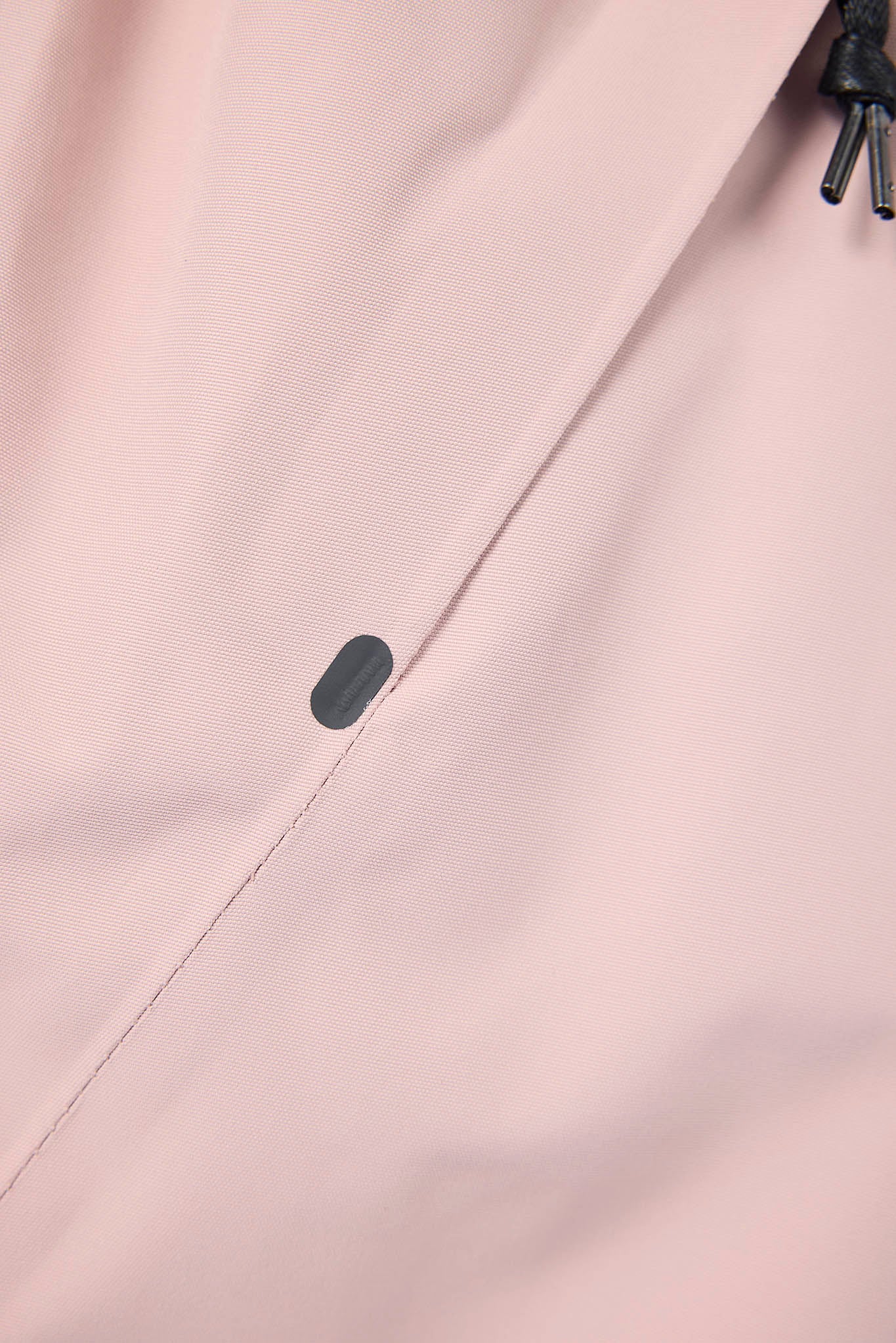 Close-up of a light blush pink fabric surface with a small dark gray oval detail and a partial black drawcord visible at the top right.
