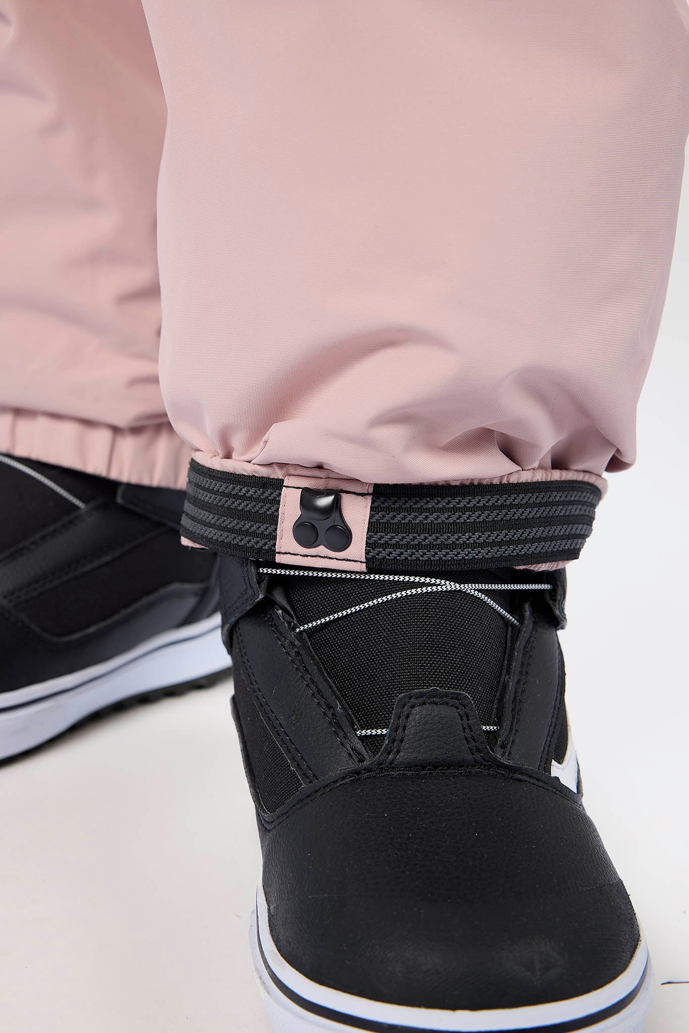 Close-up of a person wearing black snowboard boots with a black ankle strap and light pink snow pants, standing on a white surface.