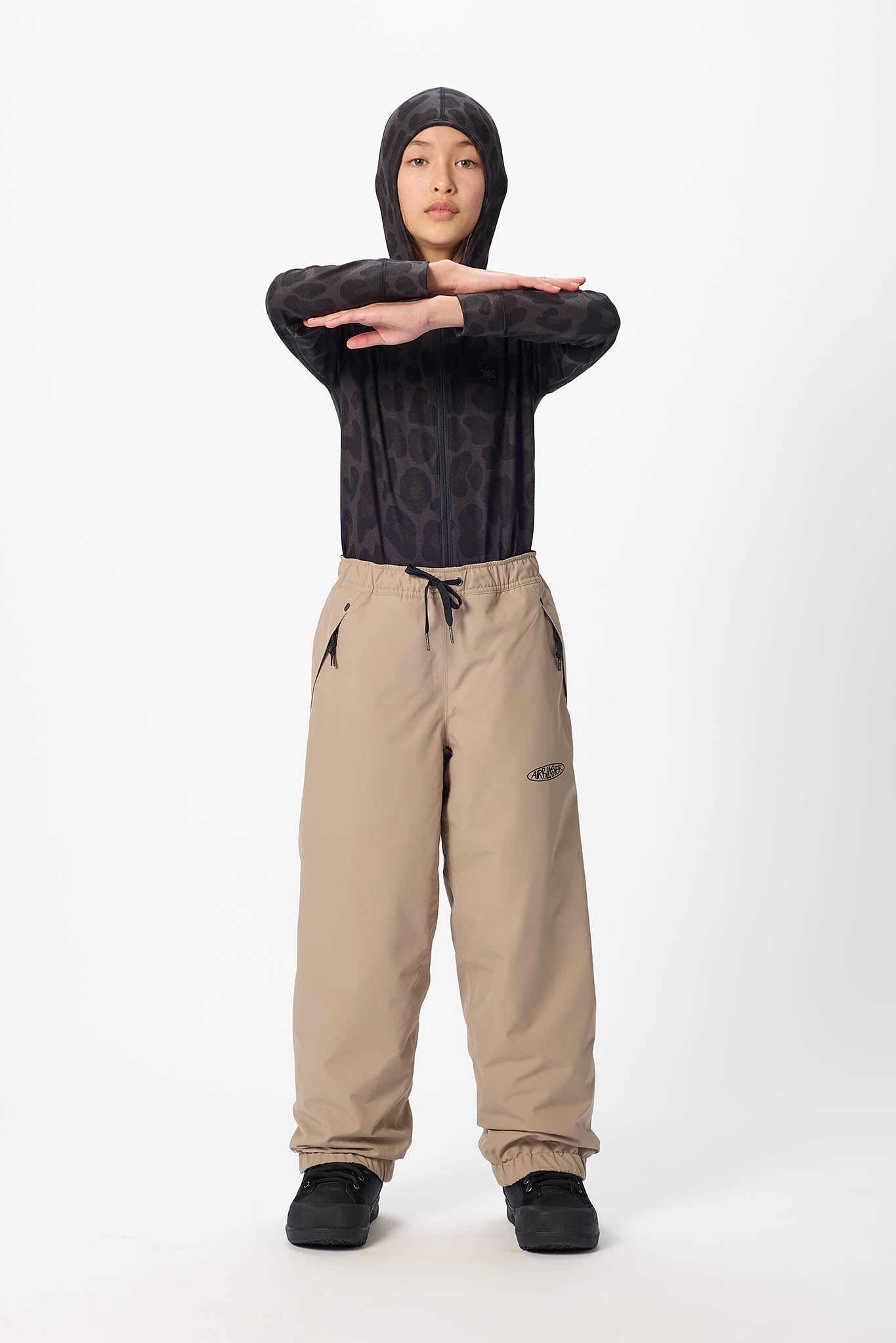 A model stands against a light gray studio background wearing a dark patterned long-sleeve hooded top, beige drawstring pants, and black shoes, with arms crossed over the chest.