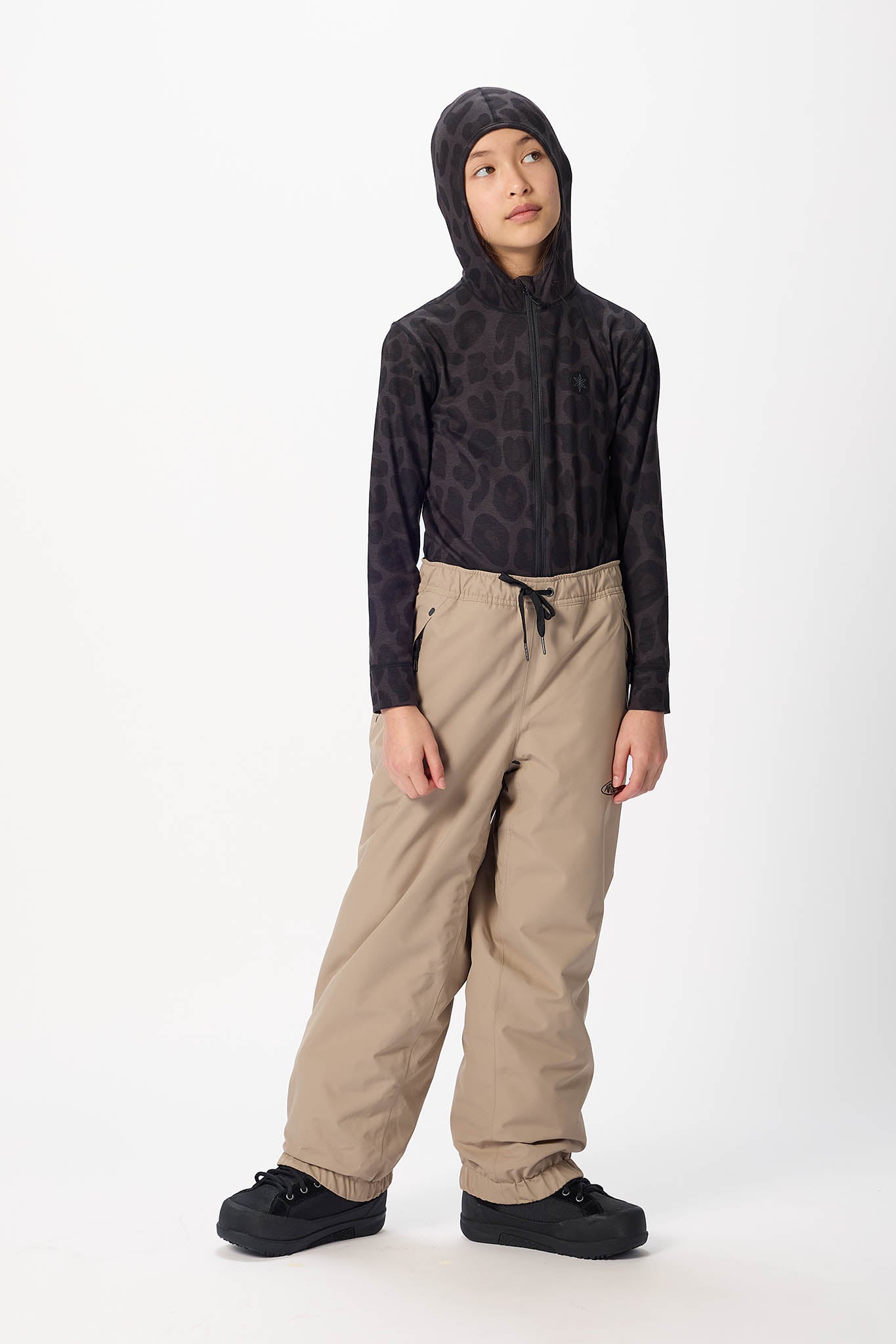 A child model wearing a black hooded long-sleeve top with a subtle tonal pattern, paired with beige drawstring snow pants and black shoes, standing against a plain white studio background.