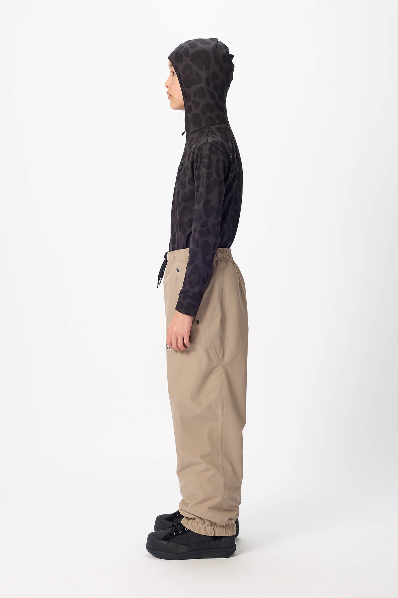 A child standing in profile wearing a dark patterned hooded sweatshirt, beige pants, and black shoes against a plain light gray studio background.