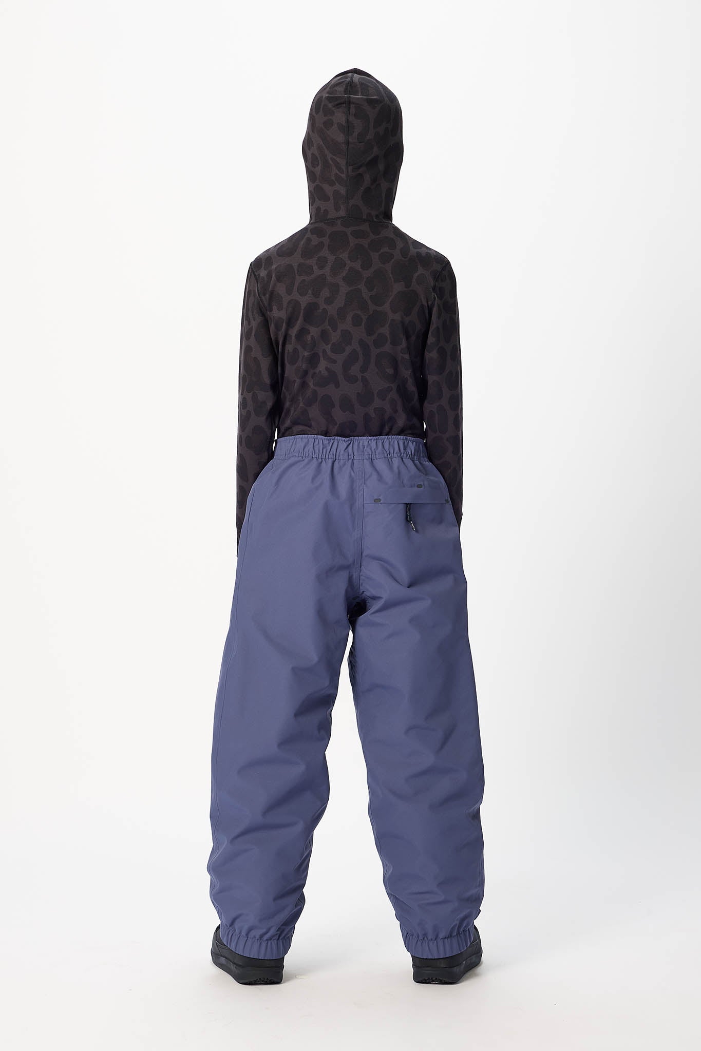 A child shown from the back wearing a dark patterned hooded sweatshirt and slate blue snow pants against a plain white studio background.