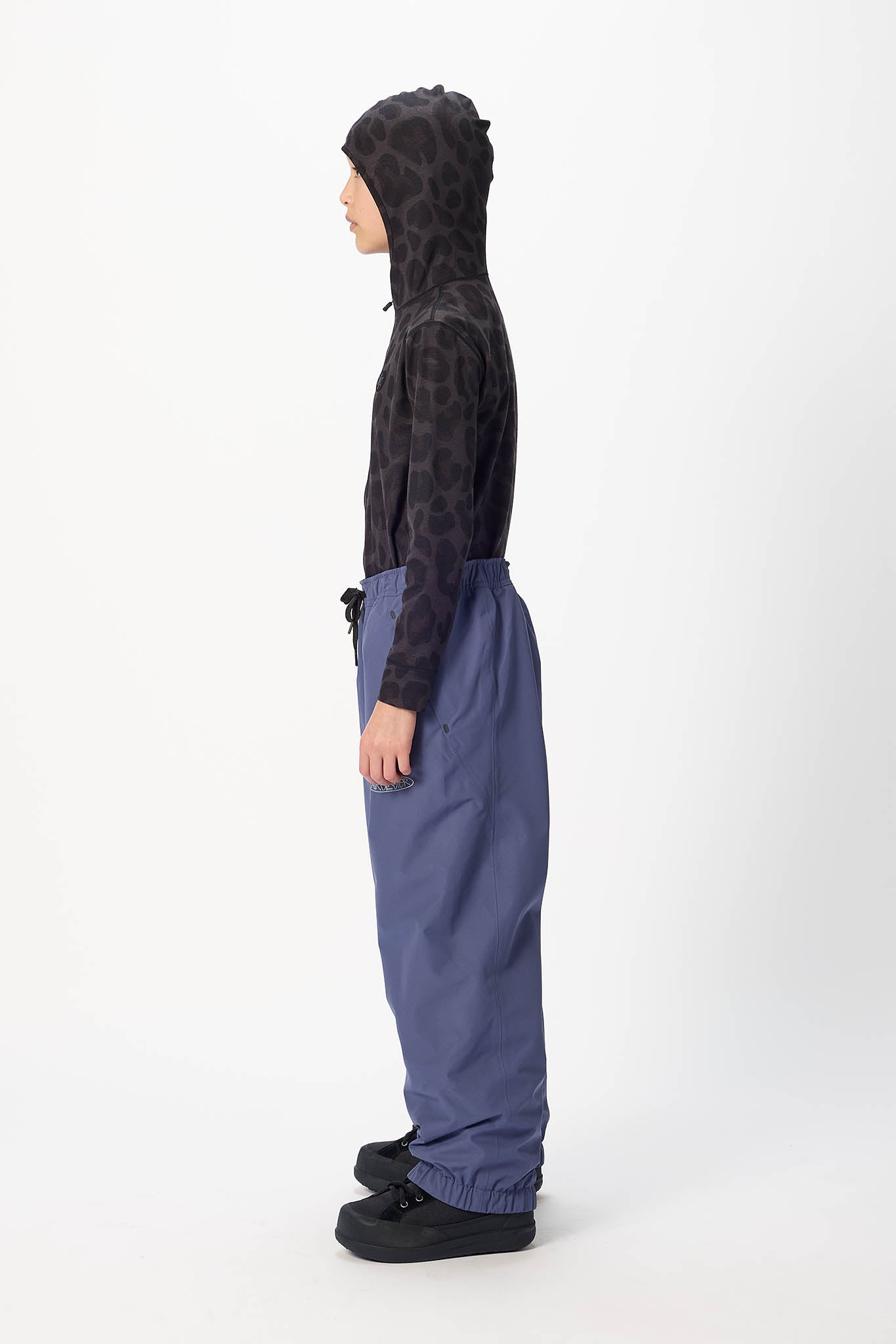 A side-profile studio shot of a model wearing a black leopard-print hooded sweatshirt and wide-leg slate blue pants against a plain white background.