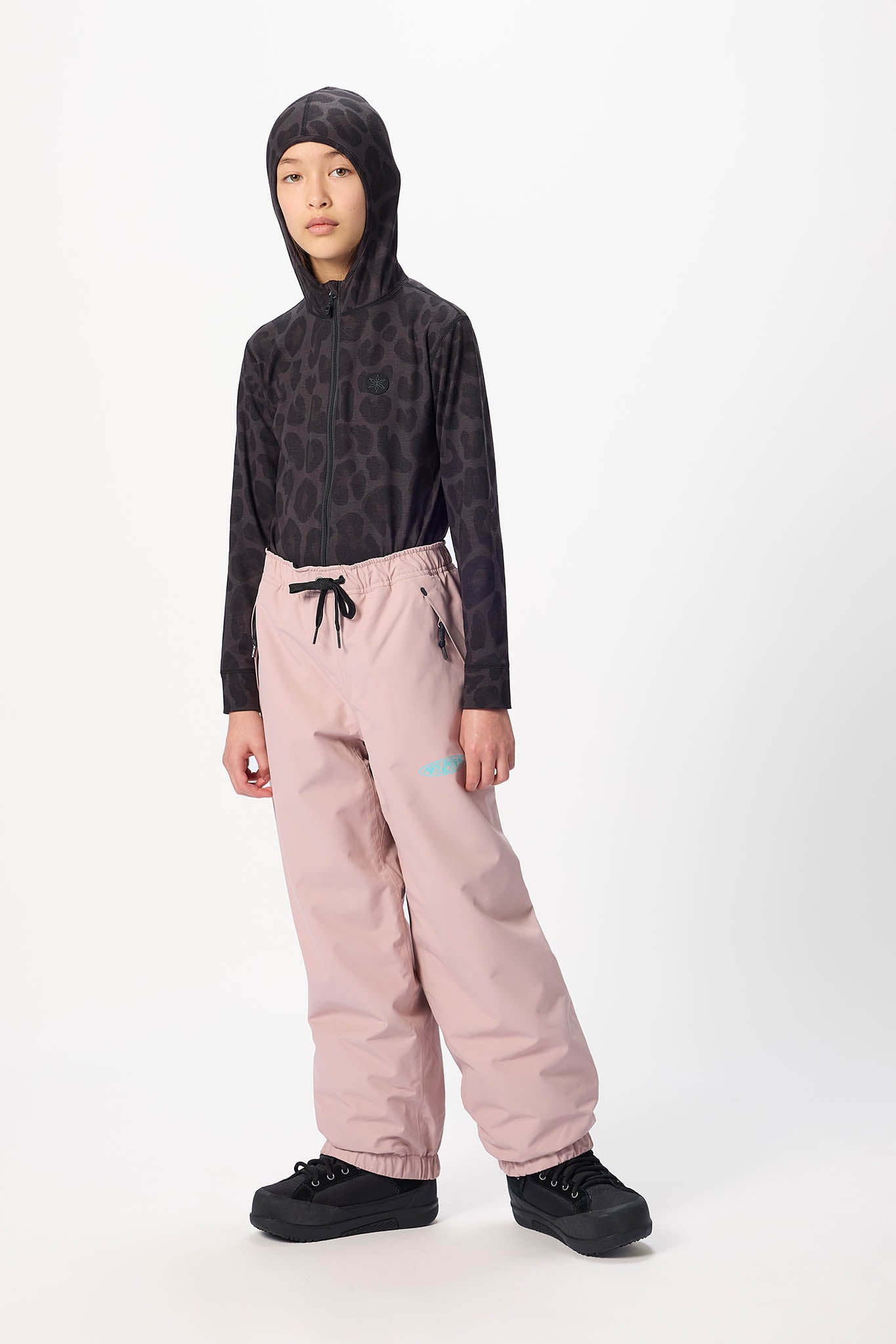 A child model wearing a black patterned long-sleeve hooded top, dusty pink snow pants with a black drawstring, and black snowboard boots, standing against a plain white studio background.