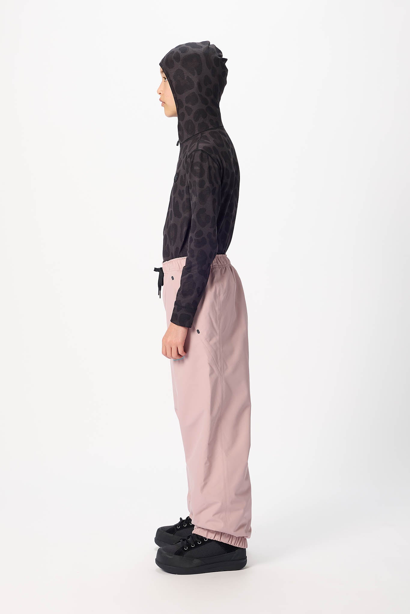 A child model shown in profile wearing a black hooded sweatshirt with a tonal animal print and light pink wide-leg pants, standing against a plain white studio background.