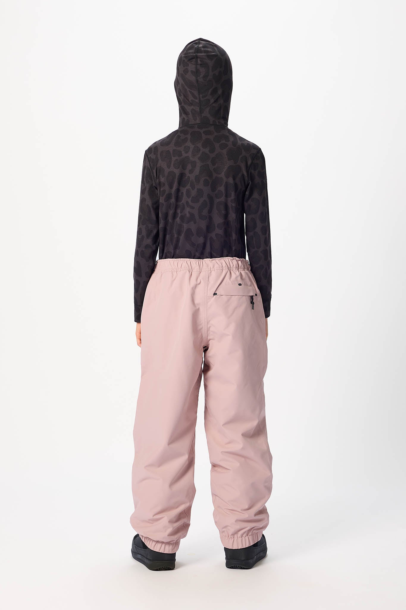 A child shown from the back wearing a black hooded long-sleeve top with an allover tonal pattern and dusty pink snow pants, standing against a plain white studio background.