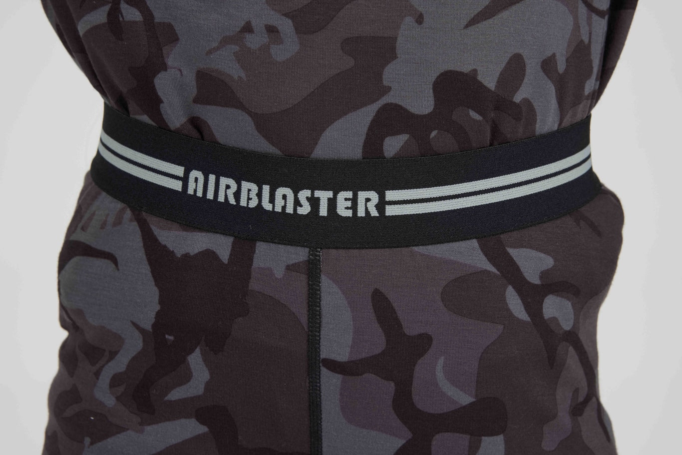 Close-up of a waistband with the brand name AIRBLASTER in white text, featuring a black elastic band with two white stripes, attached to a dark gray and black camouflage garment.