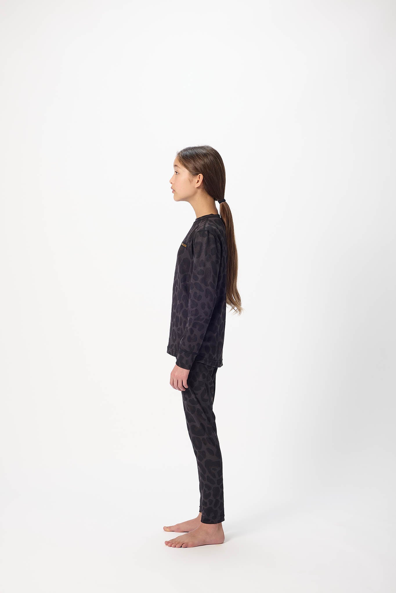 A young girl with long brown hair tied back in a ponytail stands in profile against a plain white studio background, wearing a dark patterned long-sleeve top and matching pants.