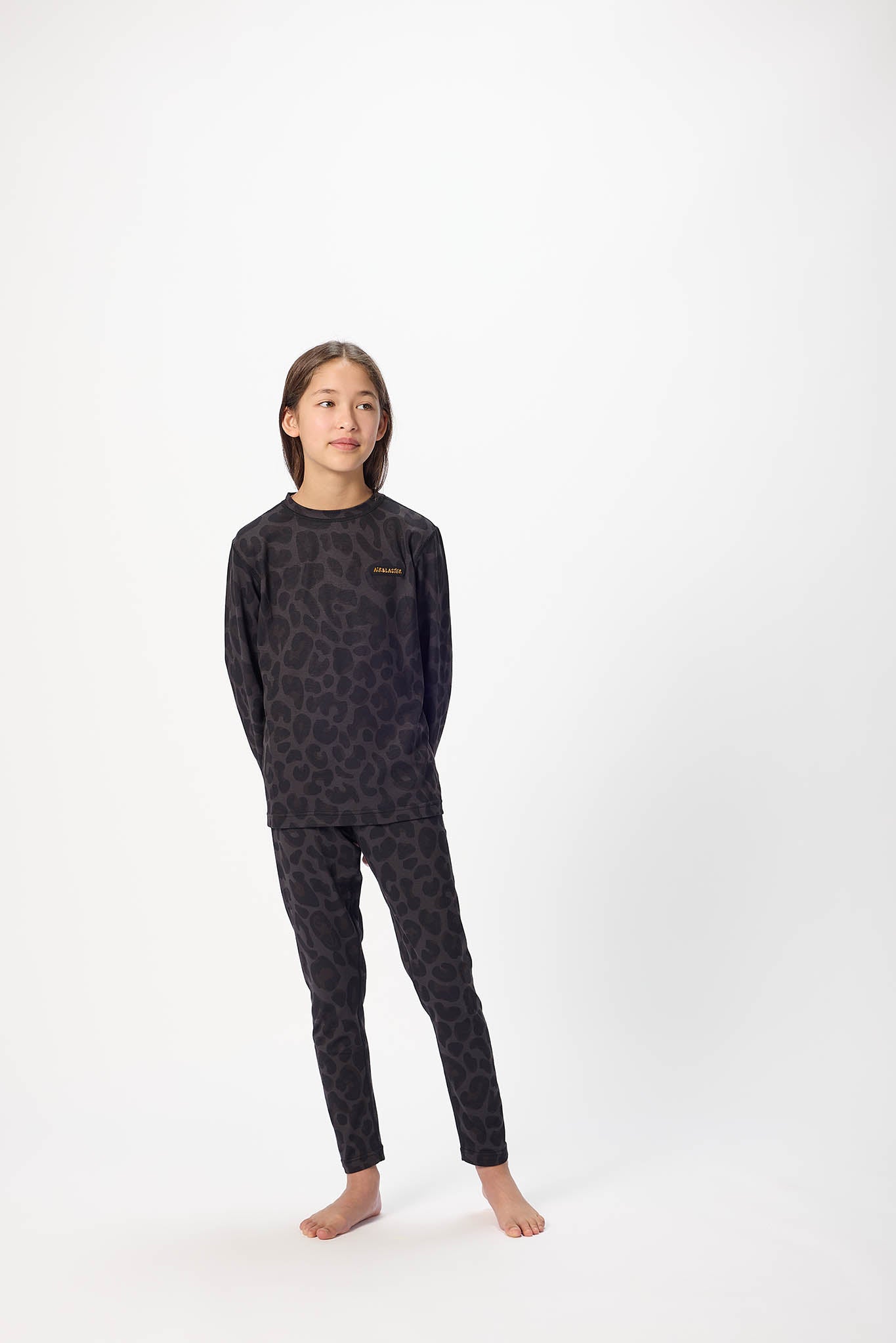 A young girl stands barefoot against a plain white studio background, wearing a dark long-sleeve pajama top and matching pants with an allover tonal animal-like pattern.