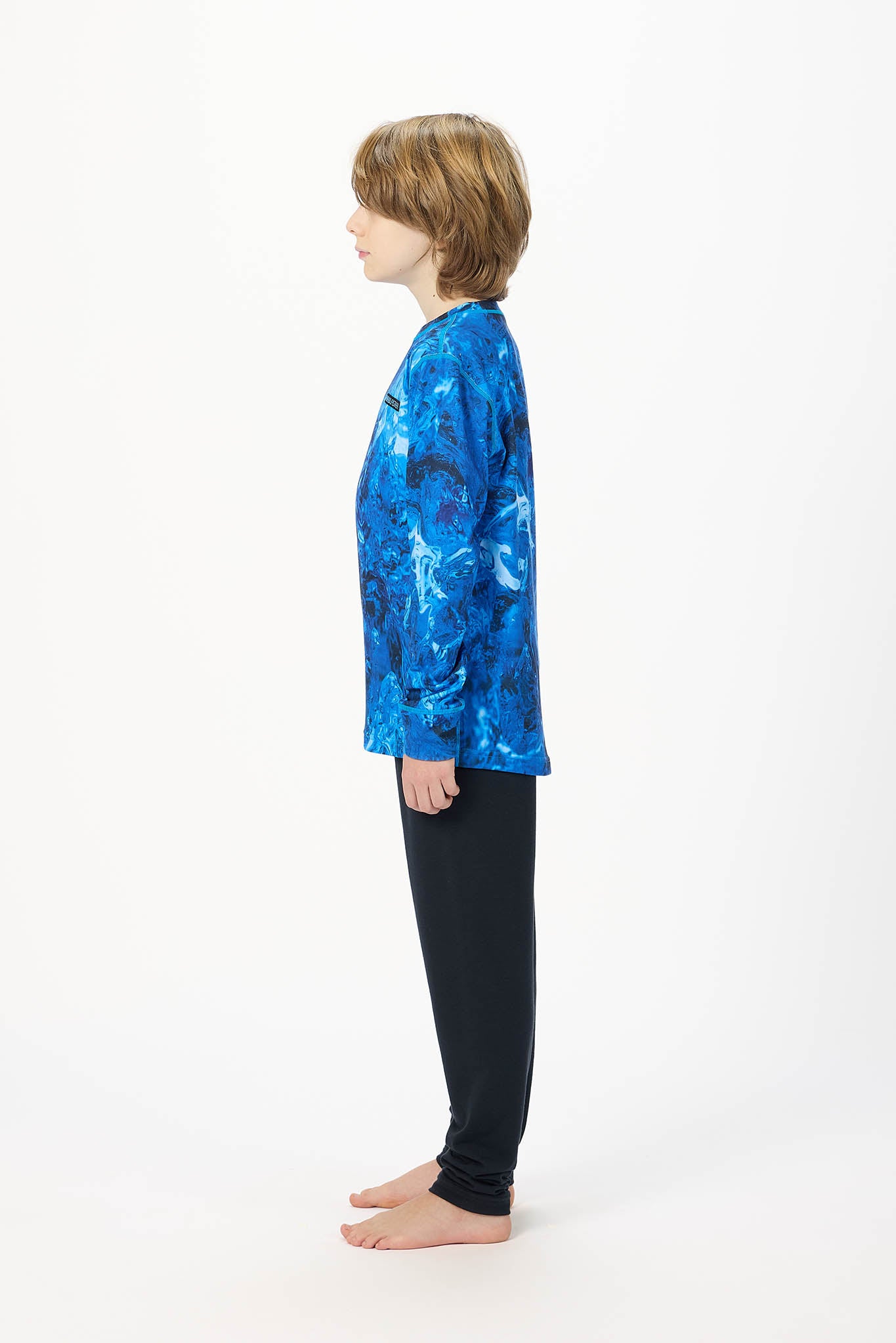 A child standing in profile wearing a long-sleeve athletic top with a blue and black abstract pattern and black pants, photographed against a plain white studio background.