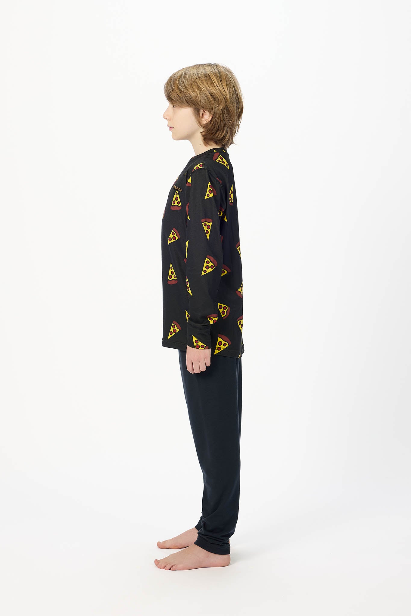 A child model shown in profile wearing a black long-sleeve pajama top with a repeating yellow triangular logo print and matching black pants, standing barefoot against a plain white studio background.
