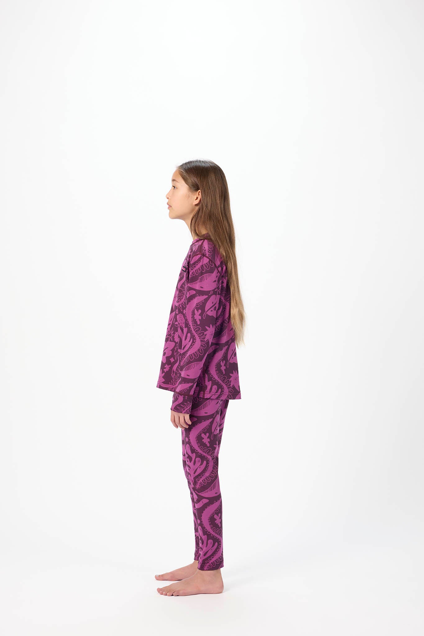 A young girl with long light brown hair stands in profile against a plain white studio background, wearing a matching two-piece pajama set with a purple and black abstract pattern.
