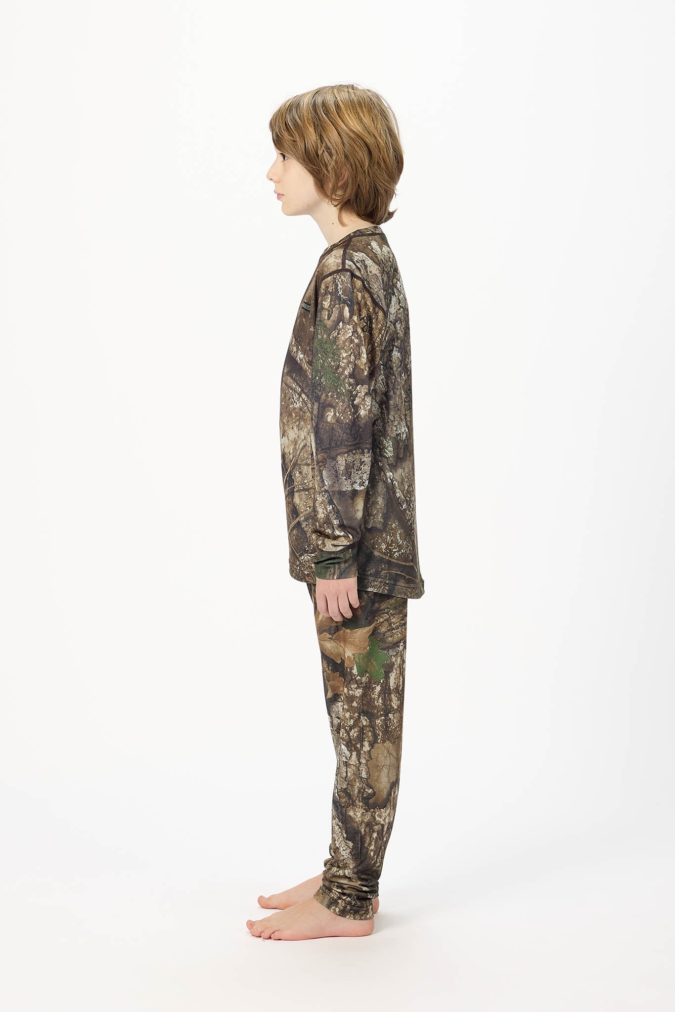 A child standing in profile wearing a camouflage long-sleeve top and matching pants against a plain white studio background.