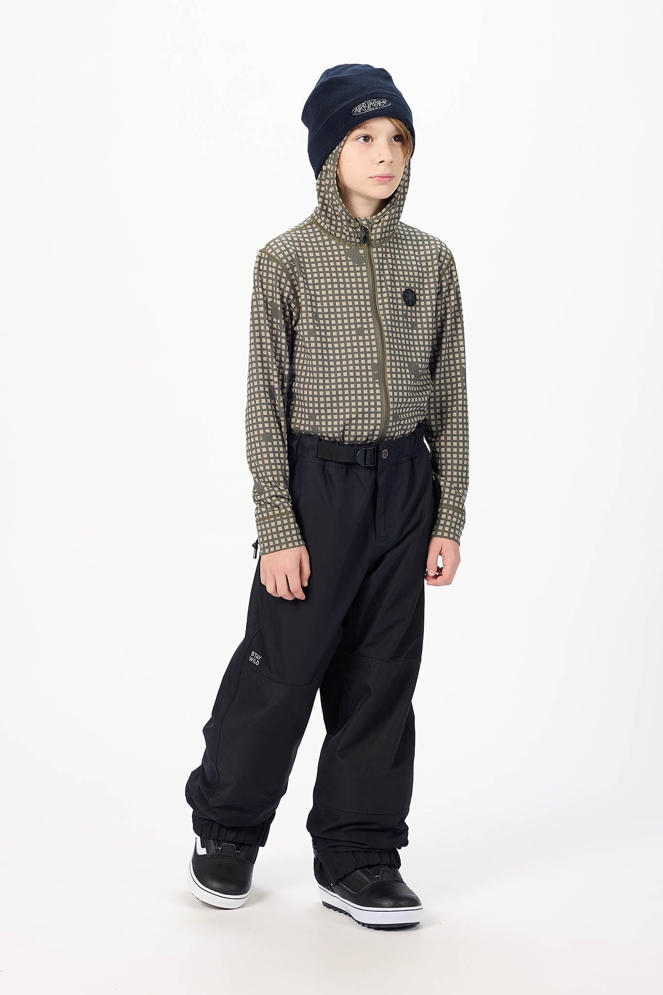 A child model wearing a patterned zip-up jacket, black snow pants, a navy beanie, and black sneakers, standing against a plain white studio background.