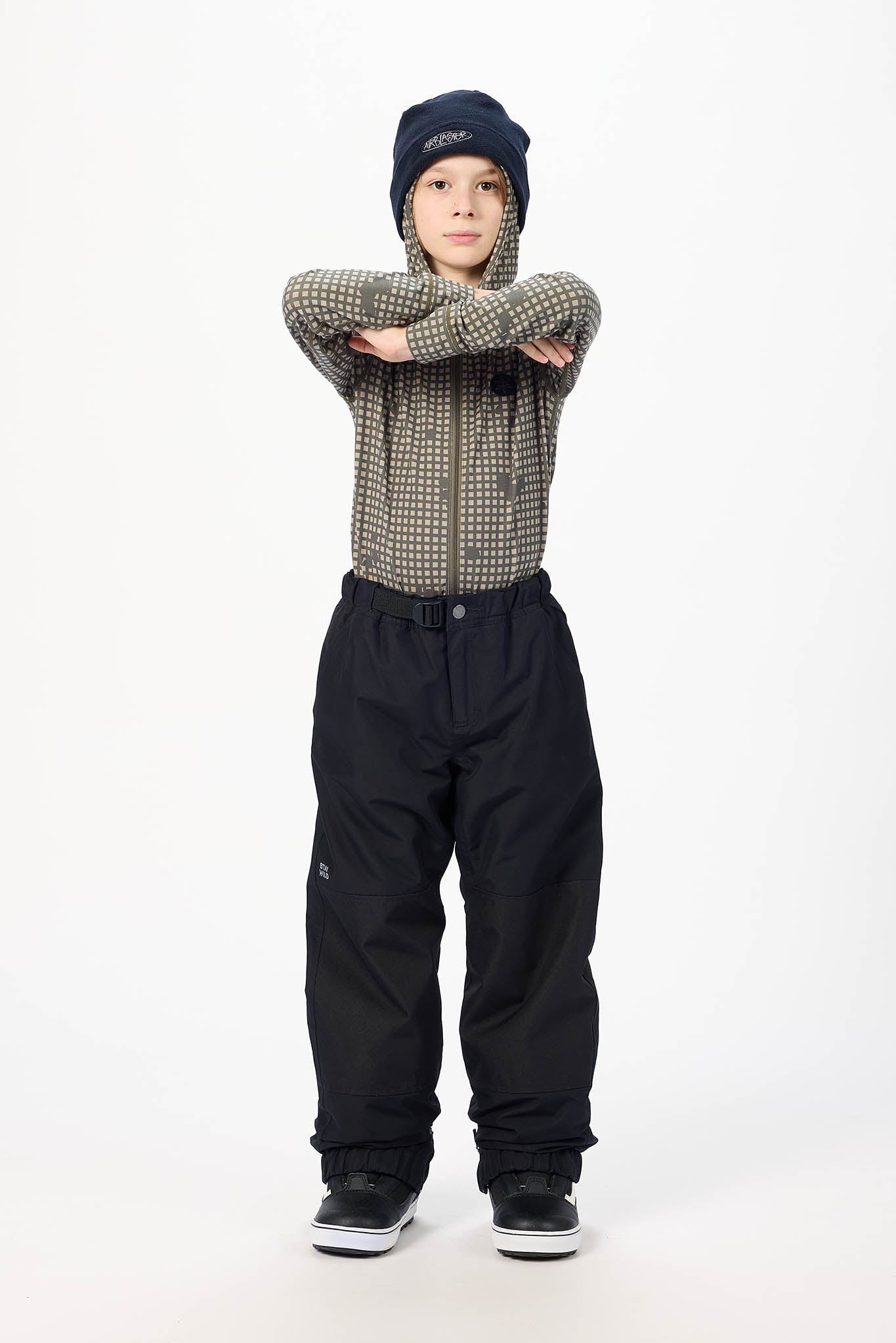 A child model wearing a dark navy beanie, a brown and black checkered long-sleeve shirt, black snow pants, and black sneakers, standing against a plain white studio background with arms crossed.