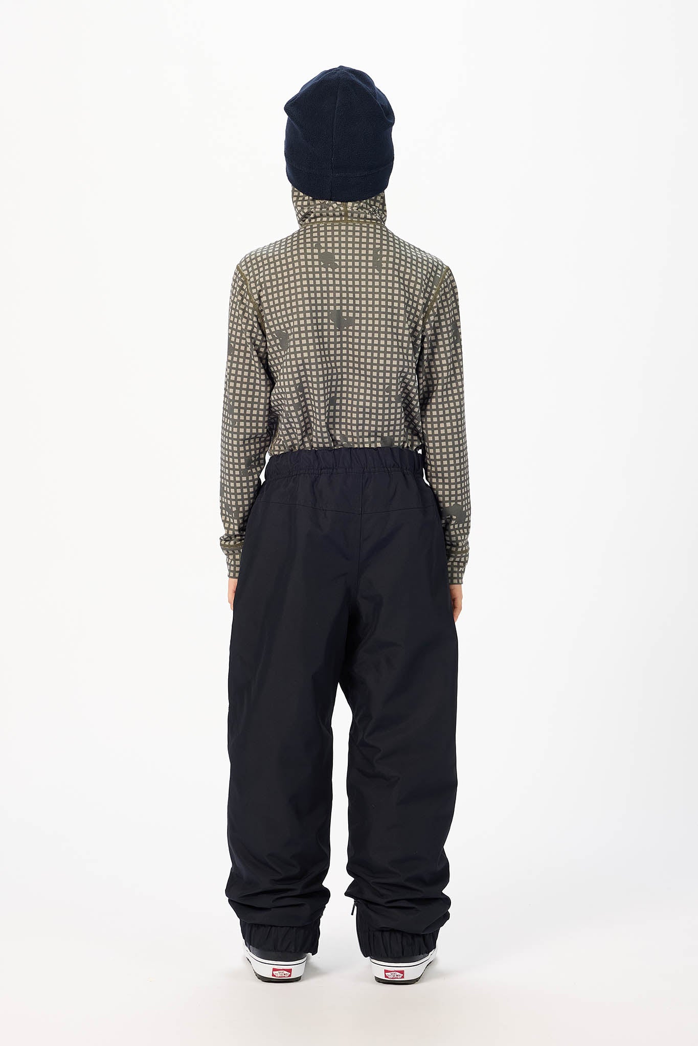 A child model shown from the back wearing a beige and black micro-check patterned long-sleeve top, black snow pants, a dark navy beanie, and white sneakers against a plain white studio background.