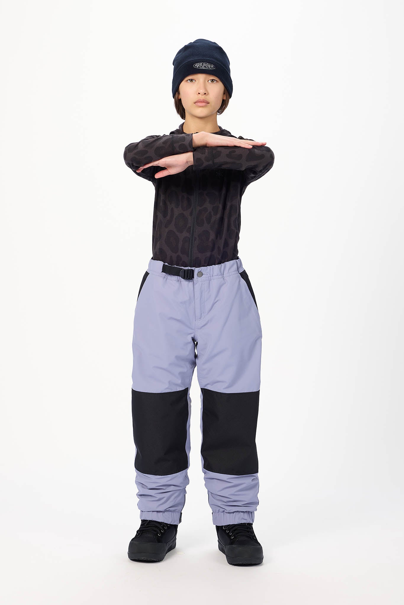 A child model wearing a dark patterned long-sleeve top, light purple and black snow pants, a dark beanie, and black shoes, standing against a plain white studio background with arms crossed.