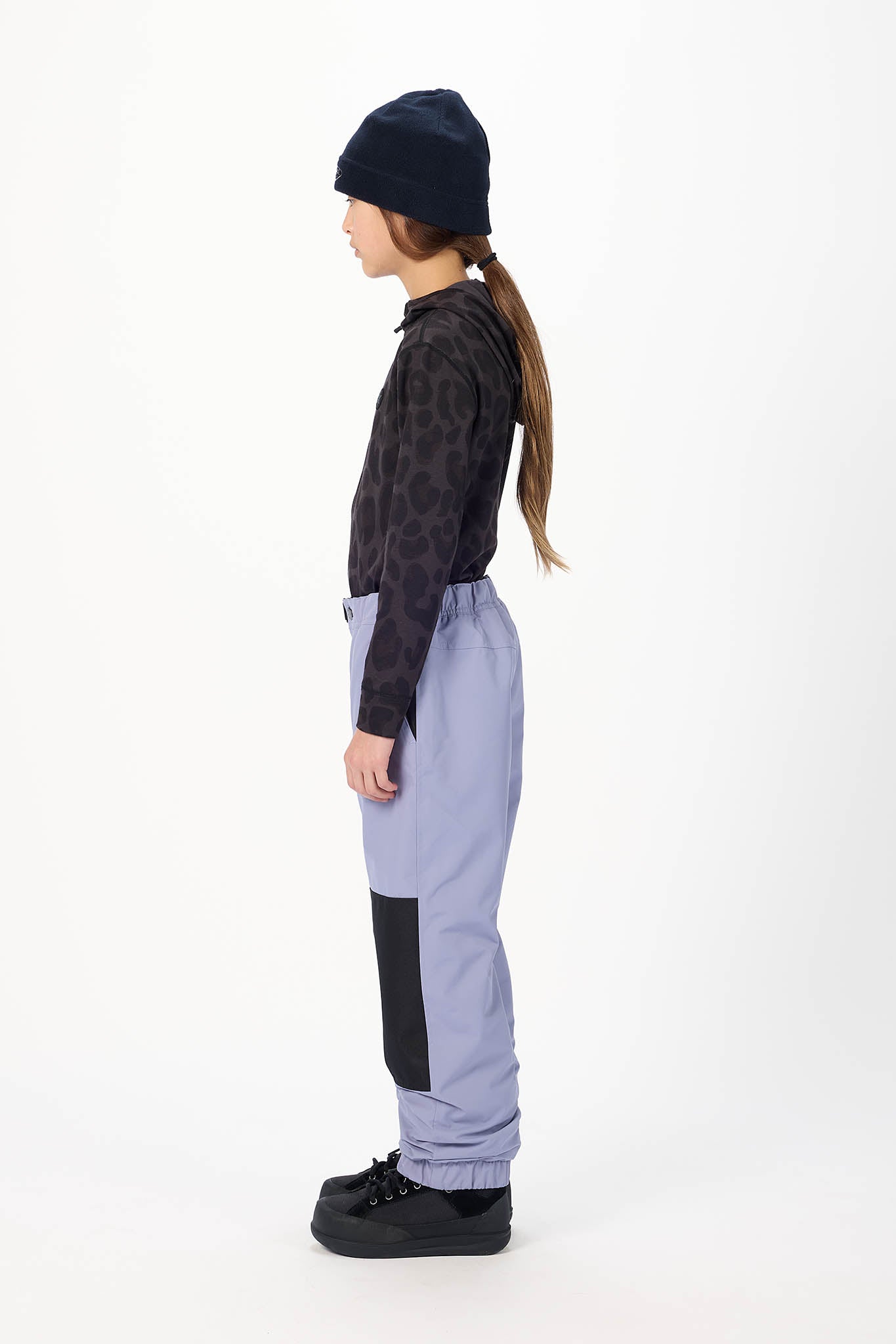 A child model shown in profile wearing a dark navy beanie, a black patterned long-sleeve top, light periwinkle blue pants with black knee patches, and black shoes, against a plain white studio background.