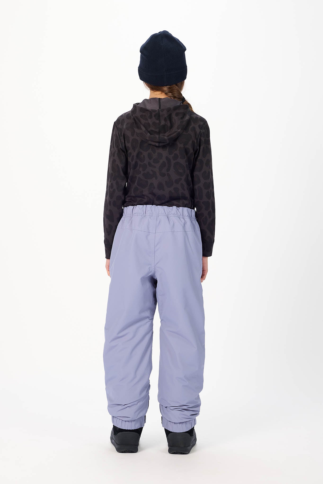 A child shown from the back wearing a dark patterned hooded sweatshirt, light purple snow pants, and a black beanie against a plain white studio background.