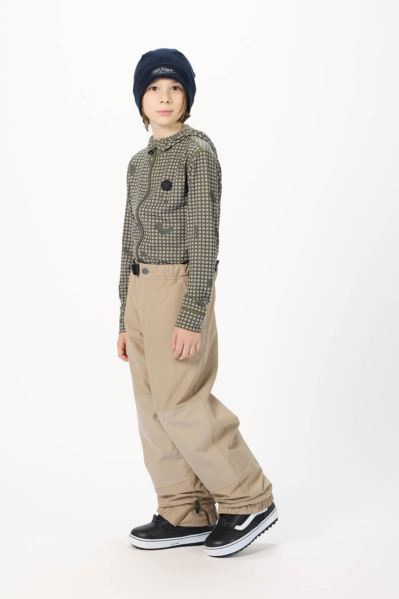 A child model wearing a dark navy beanie, a green and black checkered long-sleeve shirt, beige wide-leg pants, and black sneakers, standing against a plain white studio background.