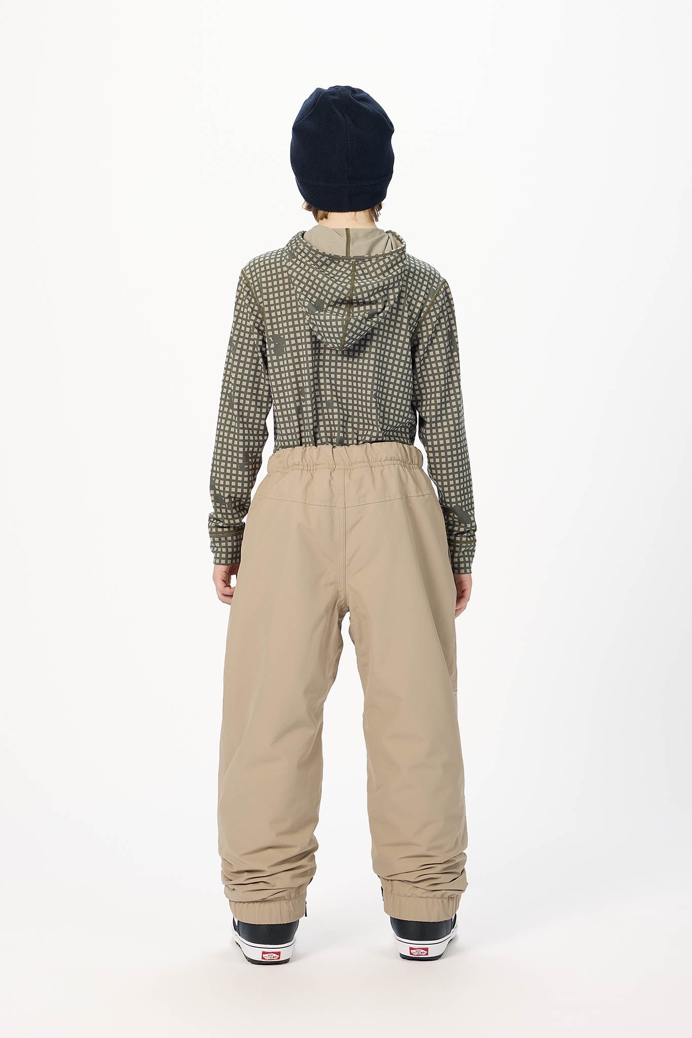 A child model shown from the back wearing a green and beige checkered long-sleeve top, beige pants, a black beanie, and black sneakers against a plain white studio background.