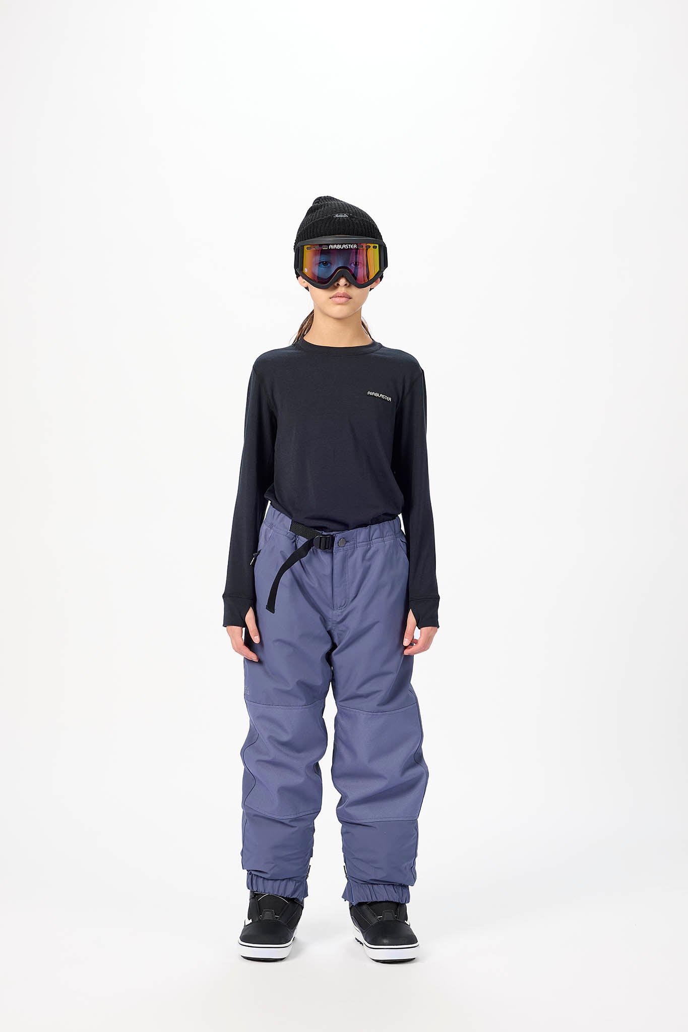 A child model wearing a black long-sleeve top, slate blue snow pants, a black beanie, and ski goggles, standing against a plain white studio background.