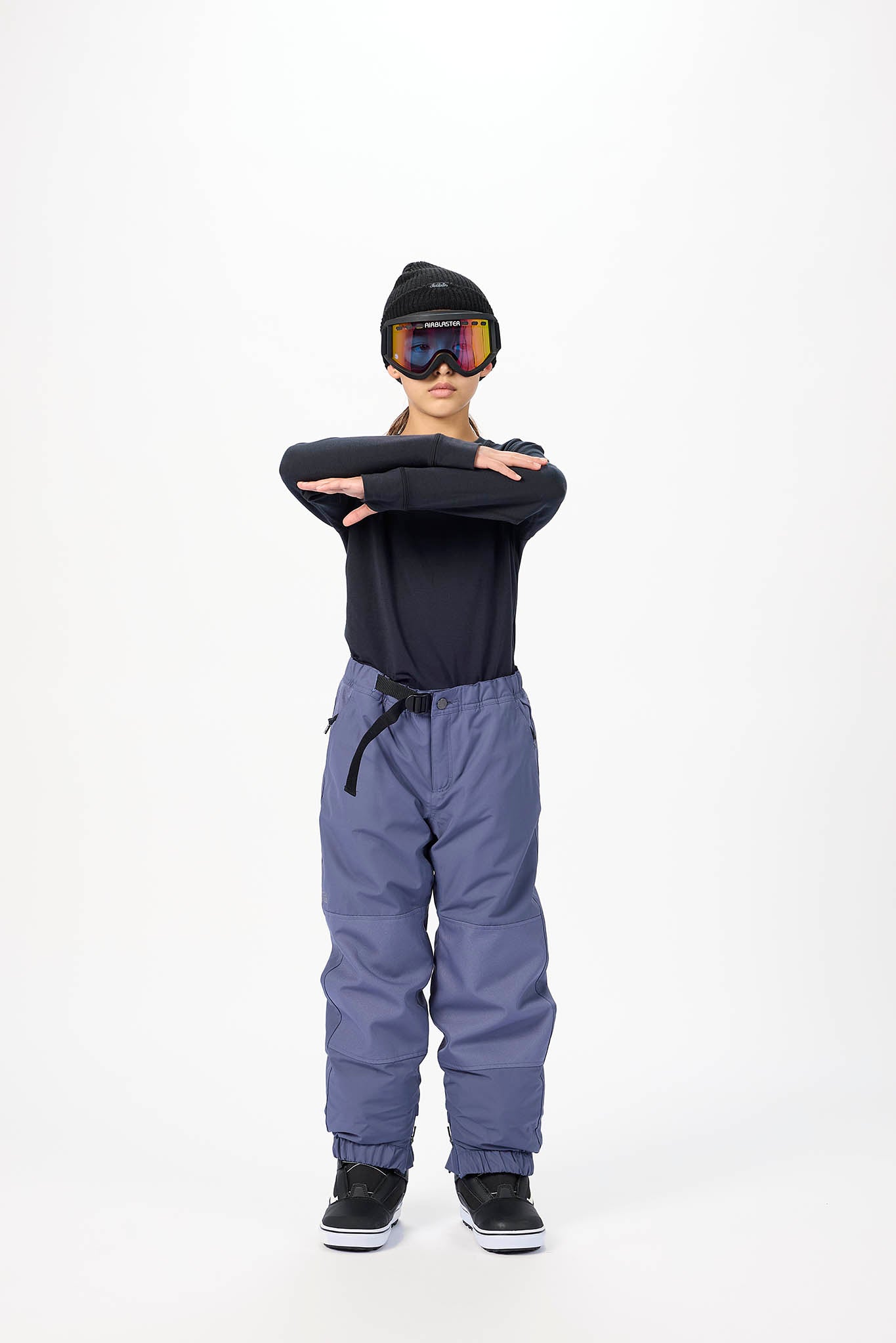 A child standing against a white studio background wearing a black beanie, ski goggles, a black long-sleeve top, and slate blue snow pants with black sneakers.