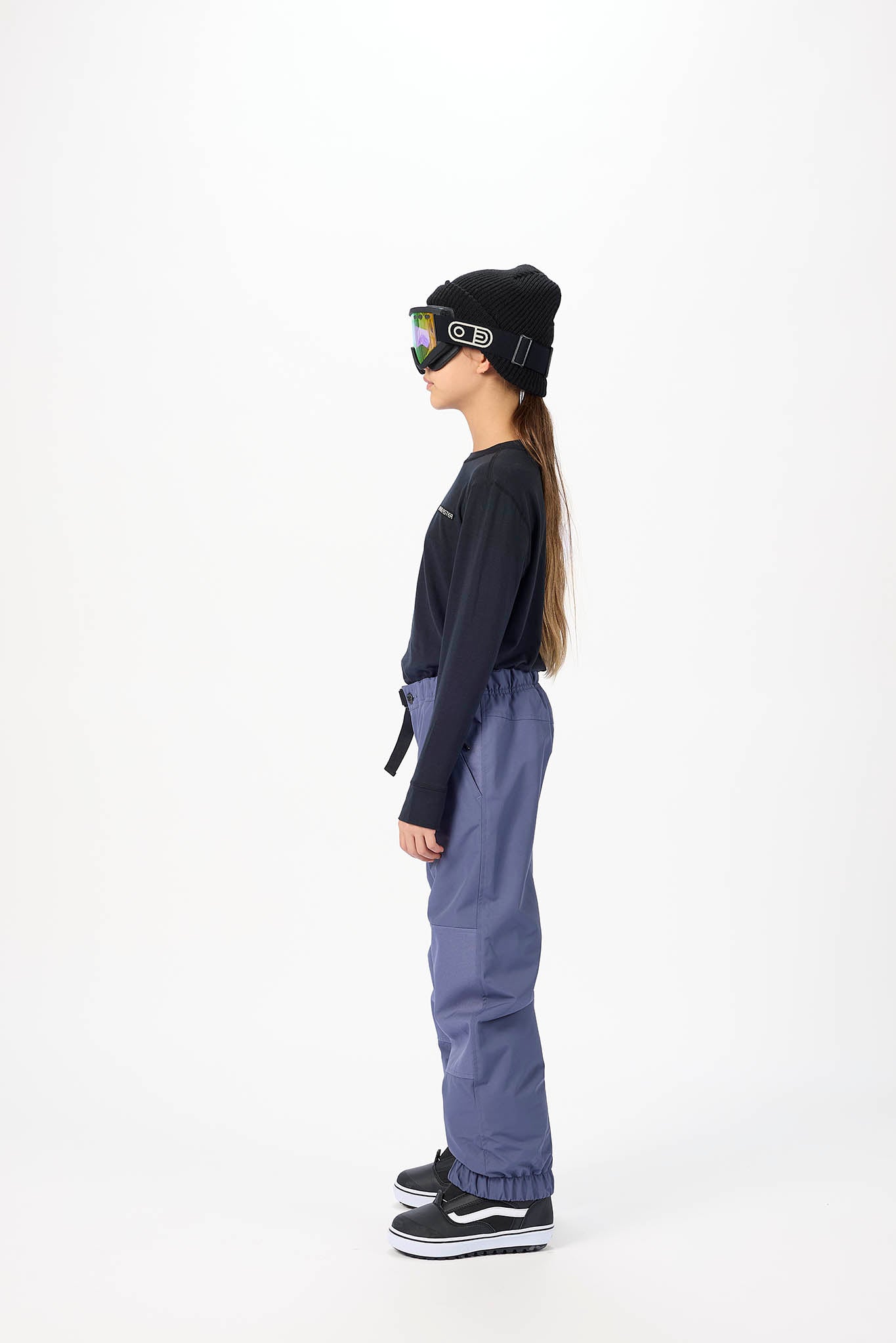 A child model shown in profile wearing a black beanie, black long-sleeve top, slate blue snow pants, and black sneakers with white soles, standing against a plain white studio background.