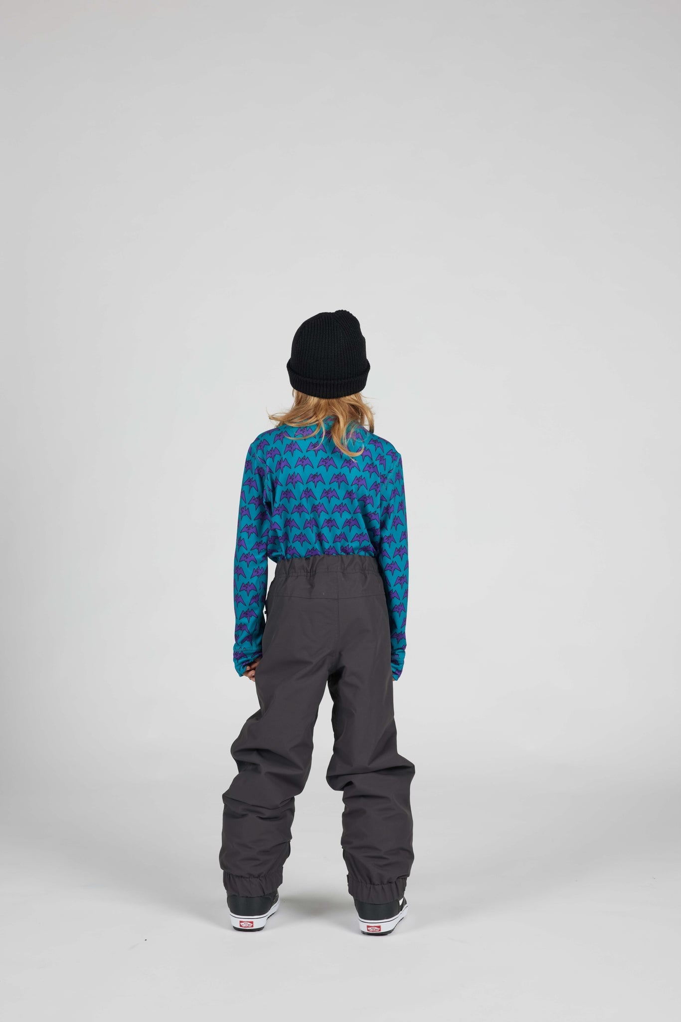 A child shown from the back wearing a black beanie, a blue and teal patterned long-sleeve top, and dark gray snow pants against a light gray studio background.