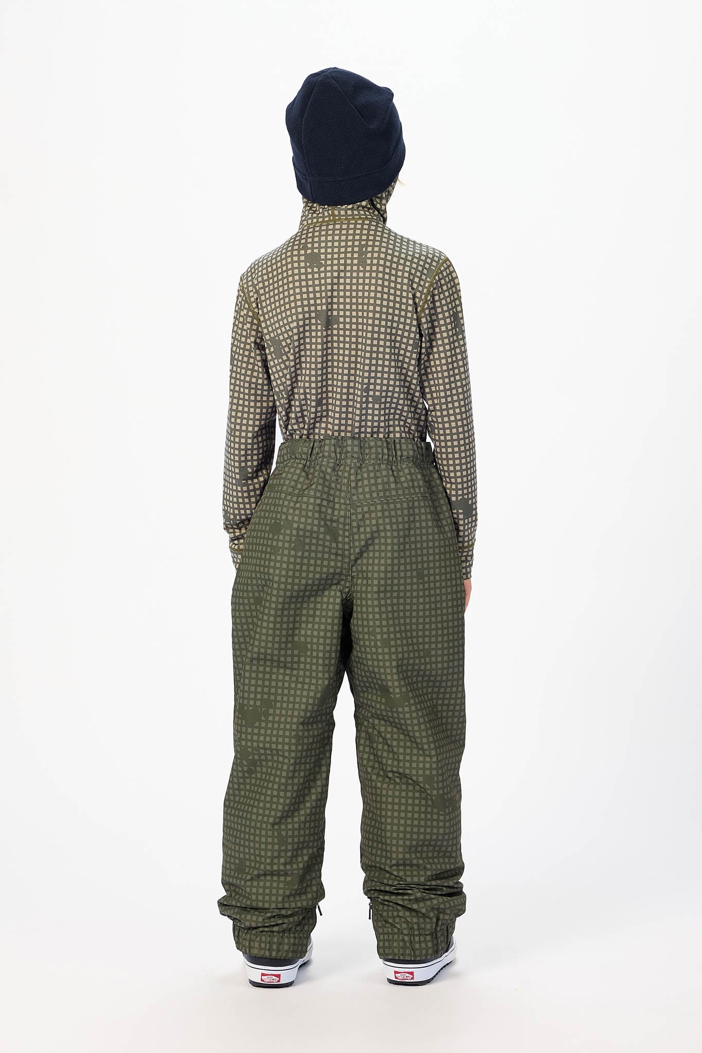 A child shown from the back wearing a dark navy knit beanie, a green and beige patterned long-sleeve top, and olive green snow pants with a subtle grid pattern, standing against a plain white studio background.