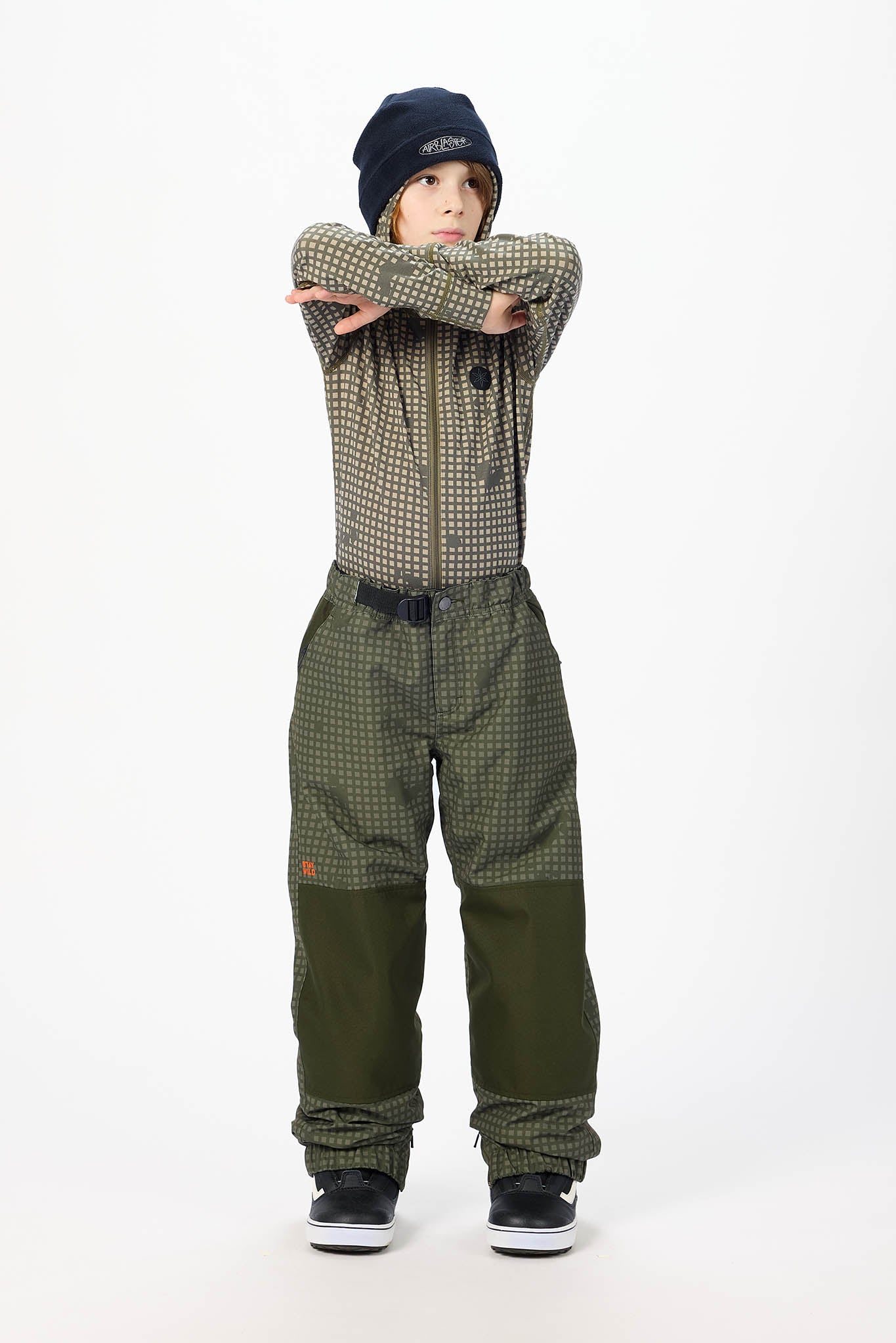 A child model wearing a green and black checkered zip-up jacket, matching green and black checkered snow pants, a dark navy beanie, and black sneakers, posing with arms crossed against a plain white studio background.
