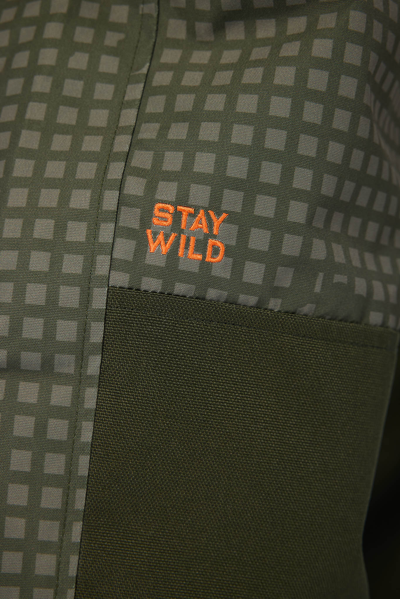 Close-up of a green and gray checkered fabric with a solid dark green pocket and orange embroidered text reading 'STAY WILD'.