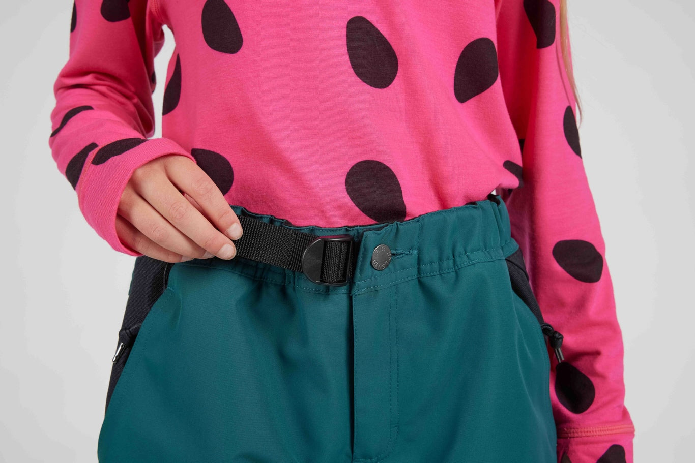 Close-up of a person wearing a bright pink long-sleeve top with large black polka dots and teal snow pants with a black waist belt.
