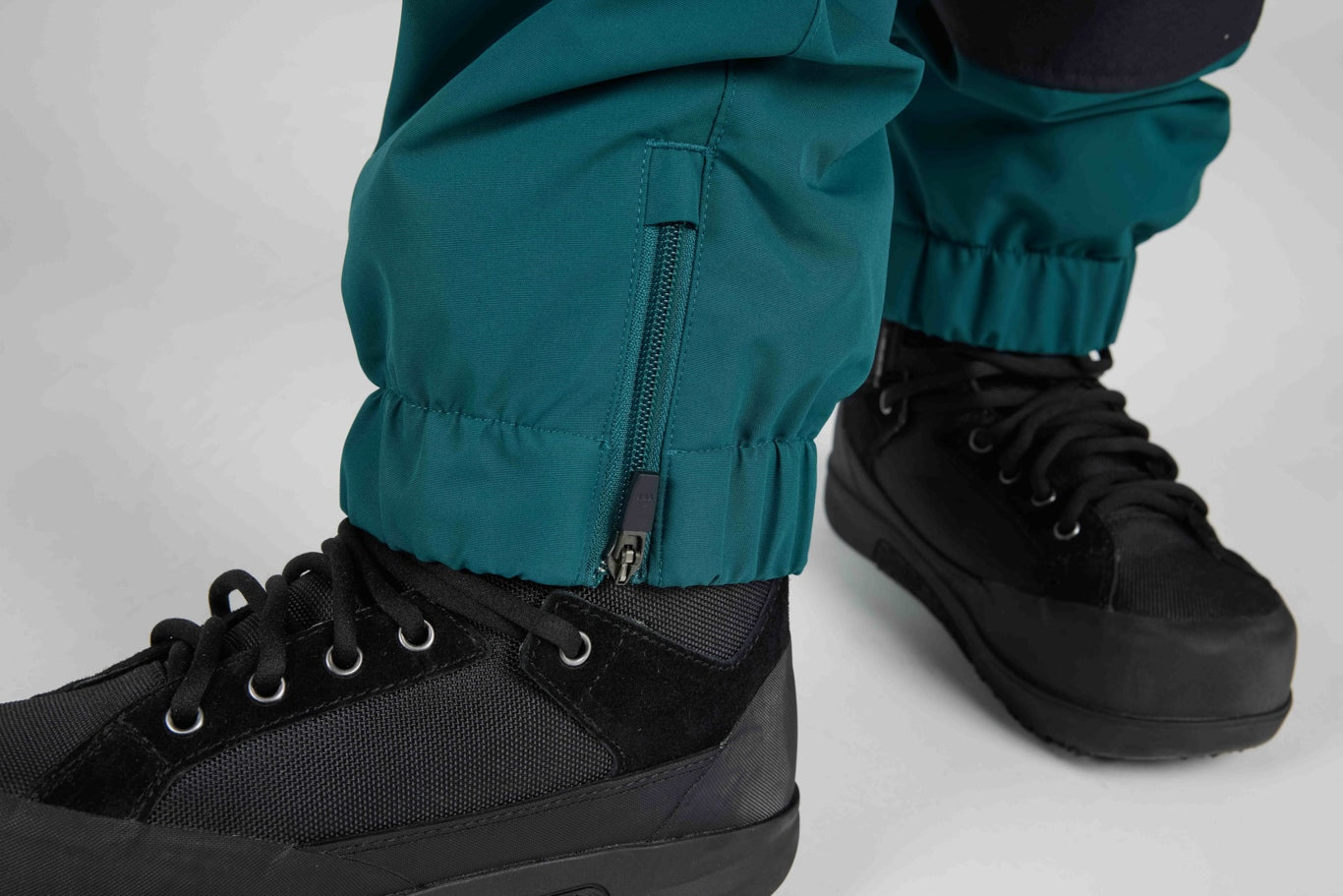 Close-up of teal pants with elastic cuffs and a zippered pocket, worn with black lace-up boots against a light gray studio background.