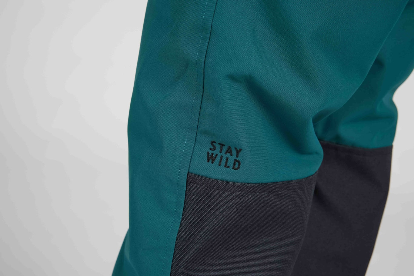 Close-up of teal and dark gray outdoor pants with the brand name 'STAY WILD' printed in black on the teal fabric.