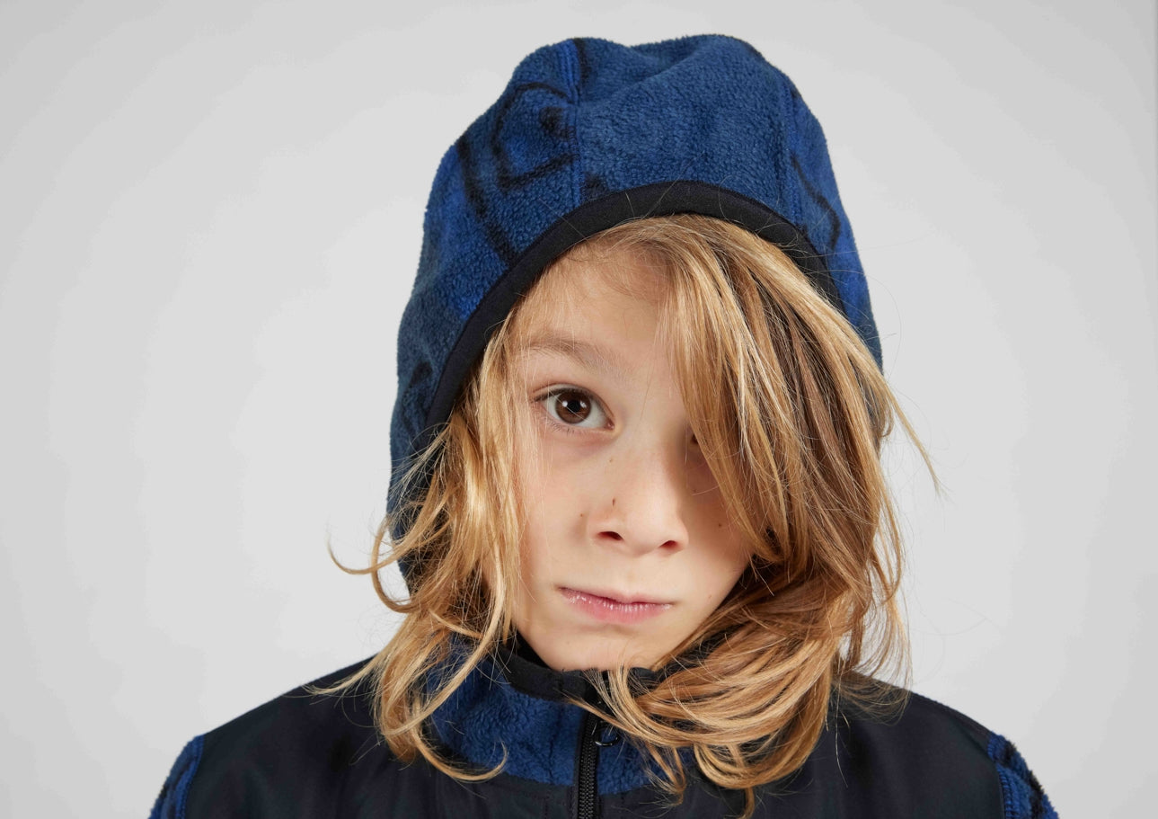 A child wearing a blue fleece hooded jacket with a matching blue patterned beanie, shown against a plain light gray studio background.