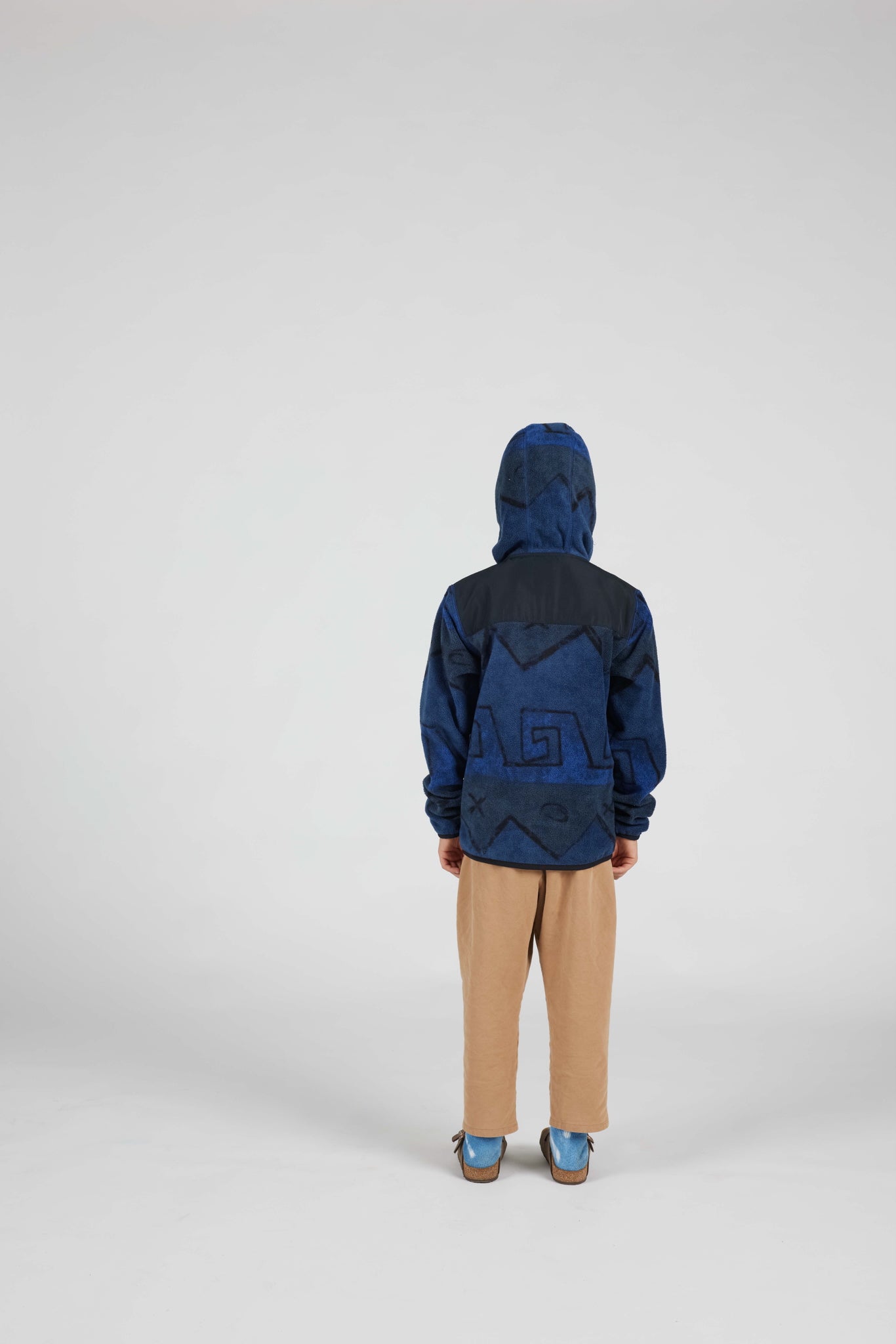 A child standing with their back to the camera, wearing a dark blue hooded jacket with a tonal geometric pattern, tan pants, and blue shoes, photographed against a neutral light gray studio background.