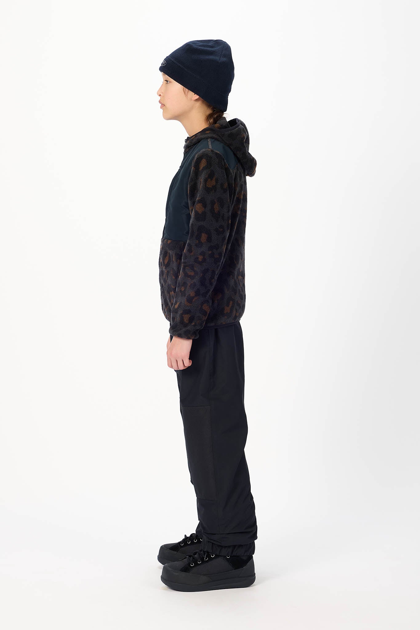 A child model shown in profile wearing a dark patterned hooded jacket, black beanie, black pants, and black shoes against a plain white studio background.
