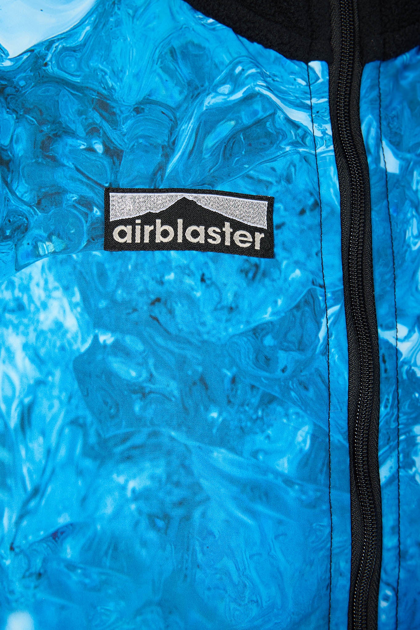 Close-up of a bright blue textured jacket with a black zipper and a black Airblaster logo patch on the chest.